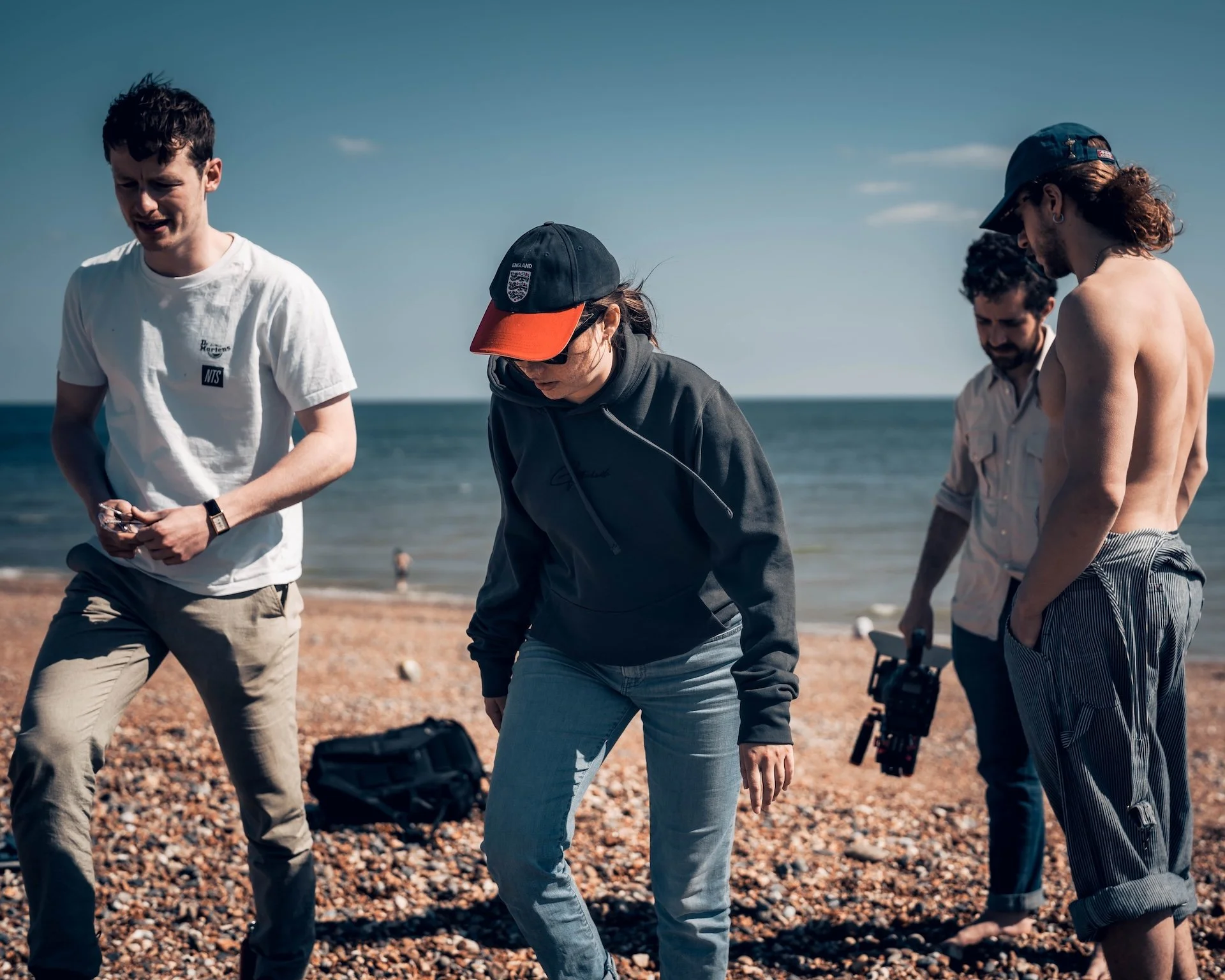 Group of five people standing on a pebble beach near the water, with one person holding a camera and others looking down or to the side, during daytime.