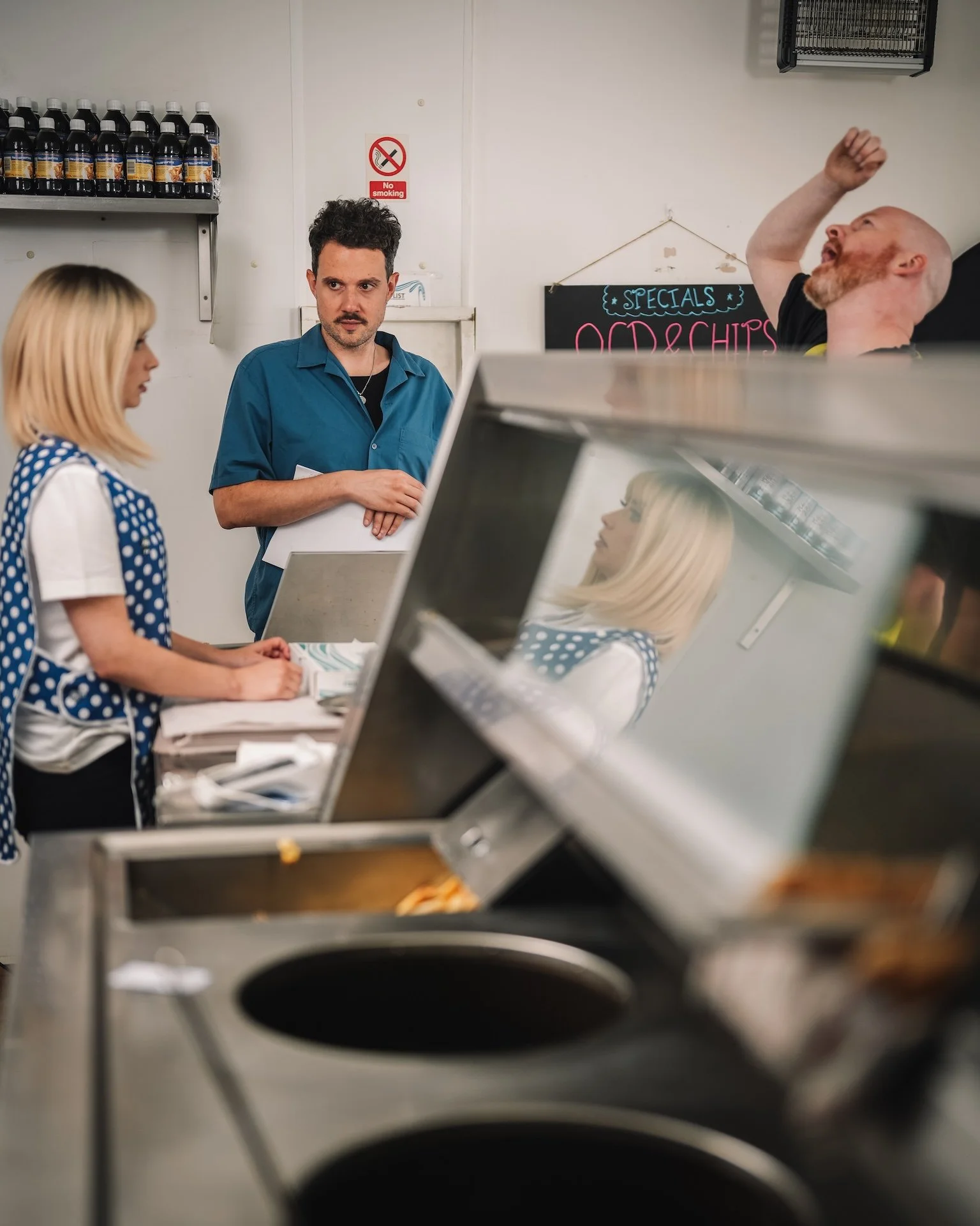 Three people stand behind a counter in a restaurant kitchen, with a chalkboard menu in the background and a shelf of bottled drinks above. One person appears to be an employee, and the other two customers, with the image partially obscured by a metal