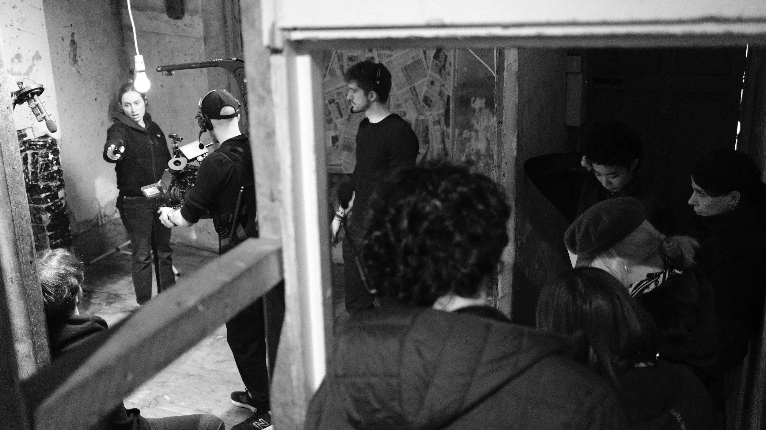 Film crew on set with a director and actors, filming scene in a rustic, dimly lit room with exposed brick and wooden beams.