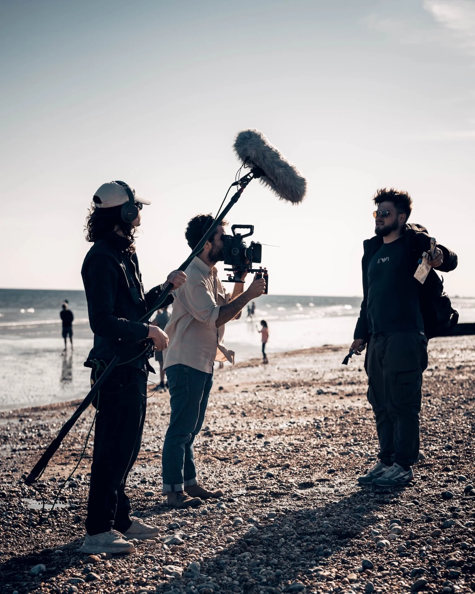 Group of people filming a man on the beach with camera crew and boom microphone, ocean in background.