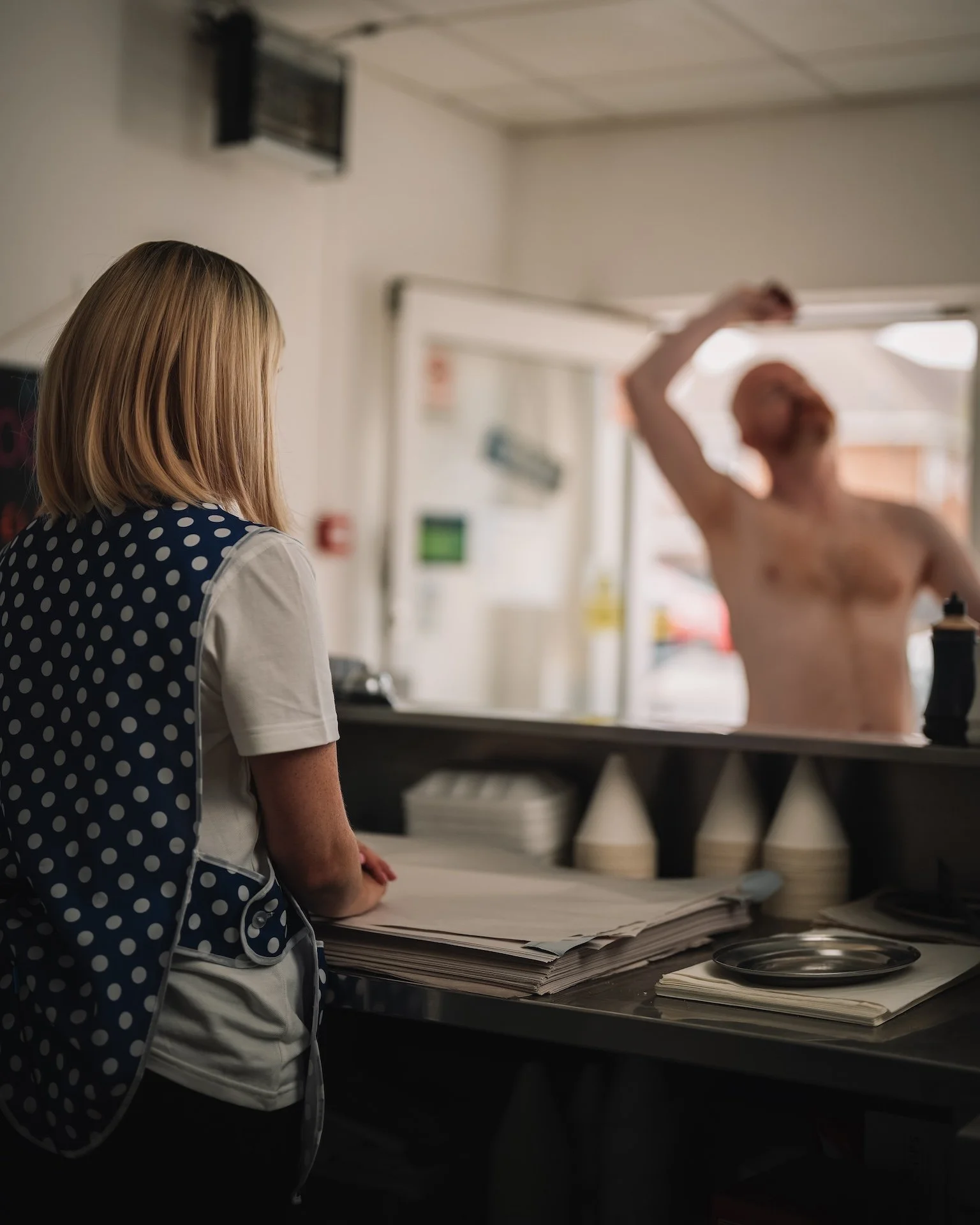 A woman with blonde hair and a polka dot apron in a kitchen or restaurant kitchen, leaning on a counter with a large stack of papers or receipts. In the background, a shirtless man with a red beard is raising his arm, possibly in a gesture or wave.