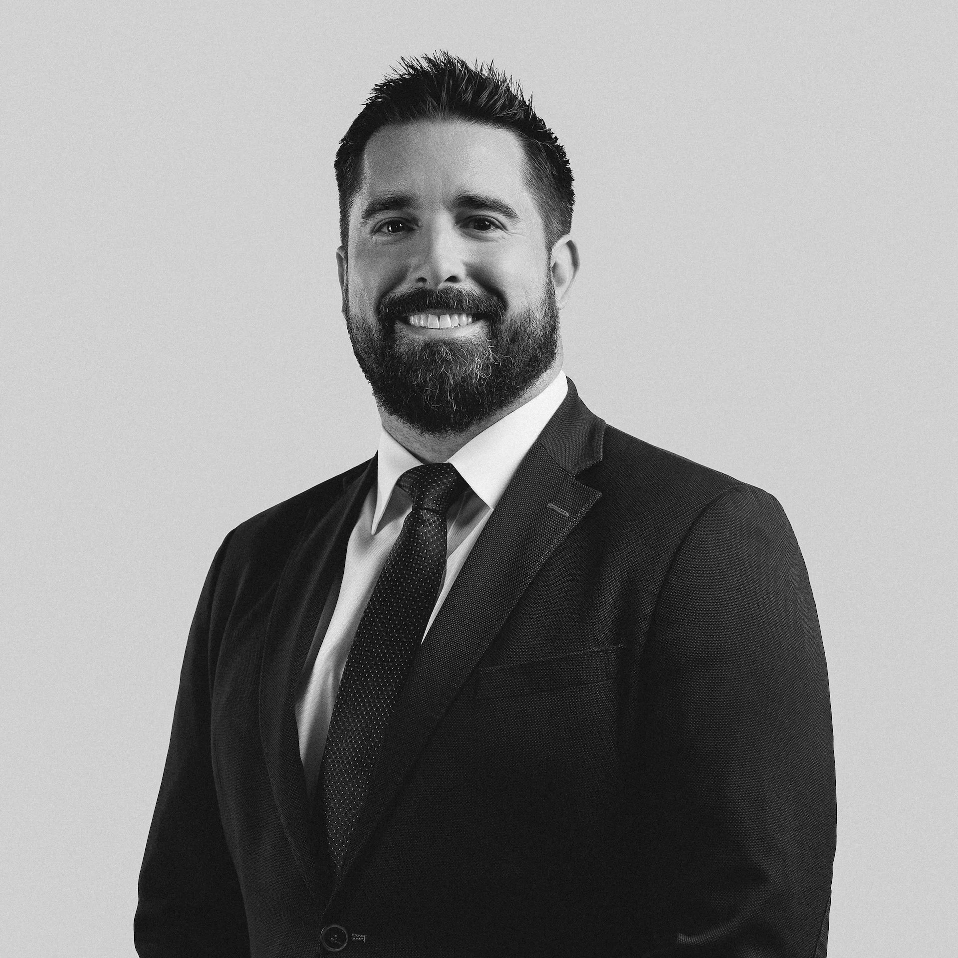 Black and white portrait of a smiling man in a suit and tie, with dark hair and a beard, against a plain background.