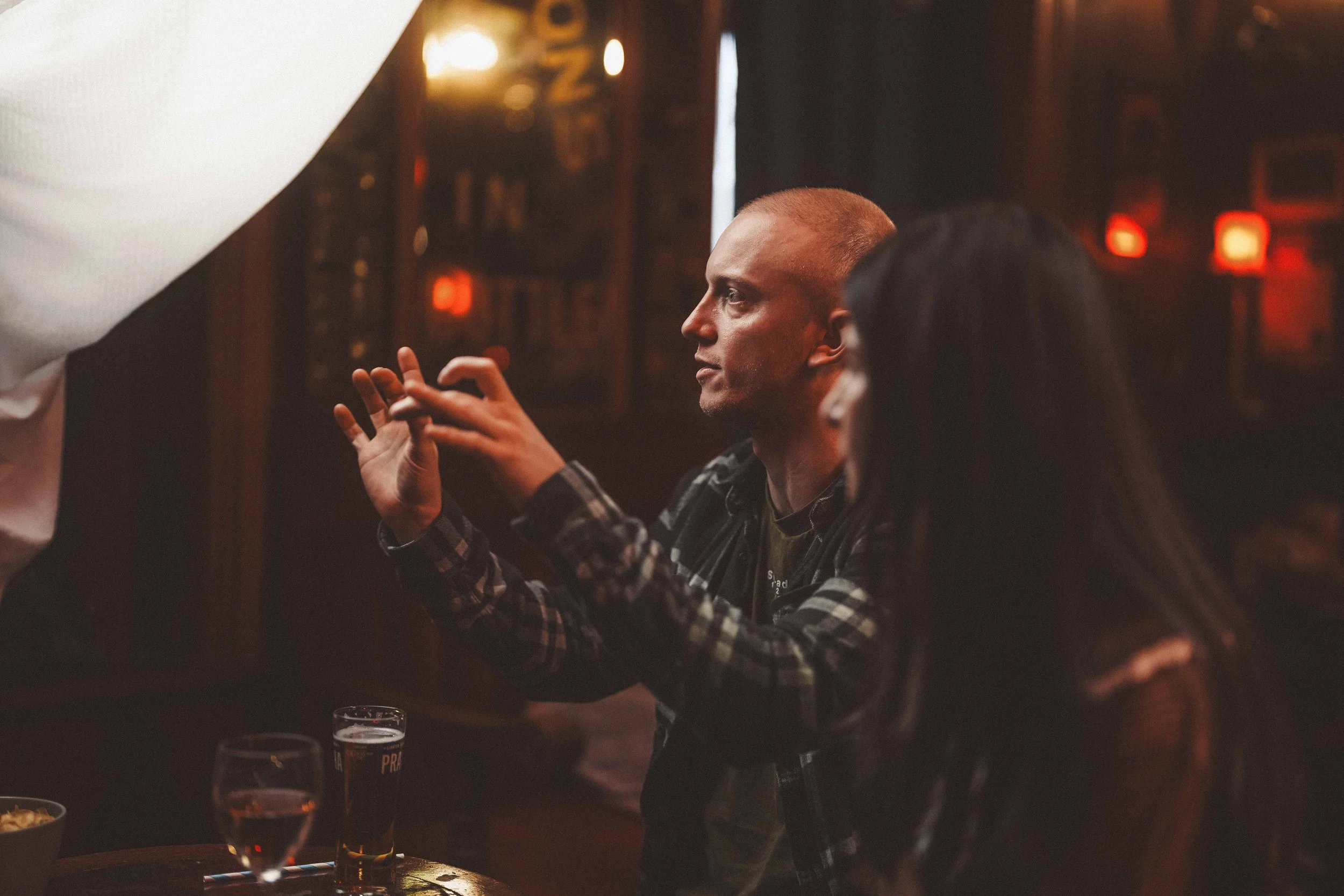 A man with a buzz cut and a plaid shirt sitting at a table in a dimly lit pub or restaurant, looking to the side with his hands clasped together. There are glasses of beer on the table and warm ambient lighting in the background.