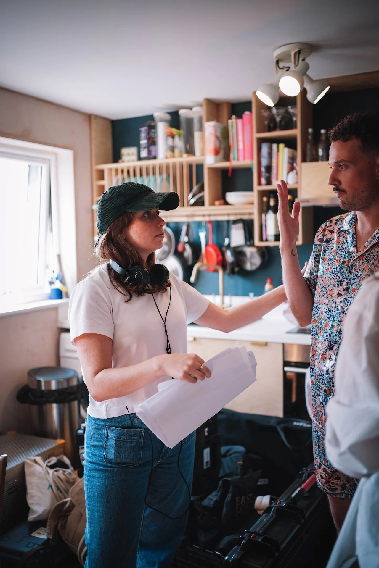 Two people in a kitchen having a conversation. One woman is wearing a green baseball cap, white t-shirt, and jeans, holding papers. The other man has short curly hair, a beard, and is gesturing with his hand. The kitchen has wooden shelves, books, an
