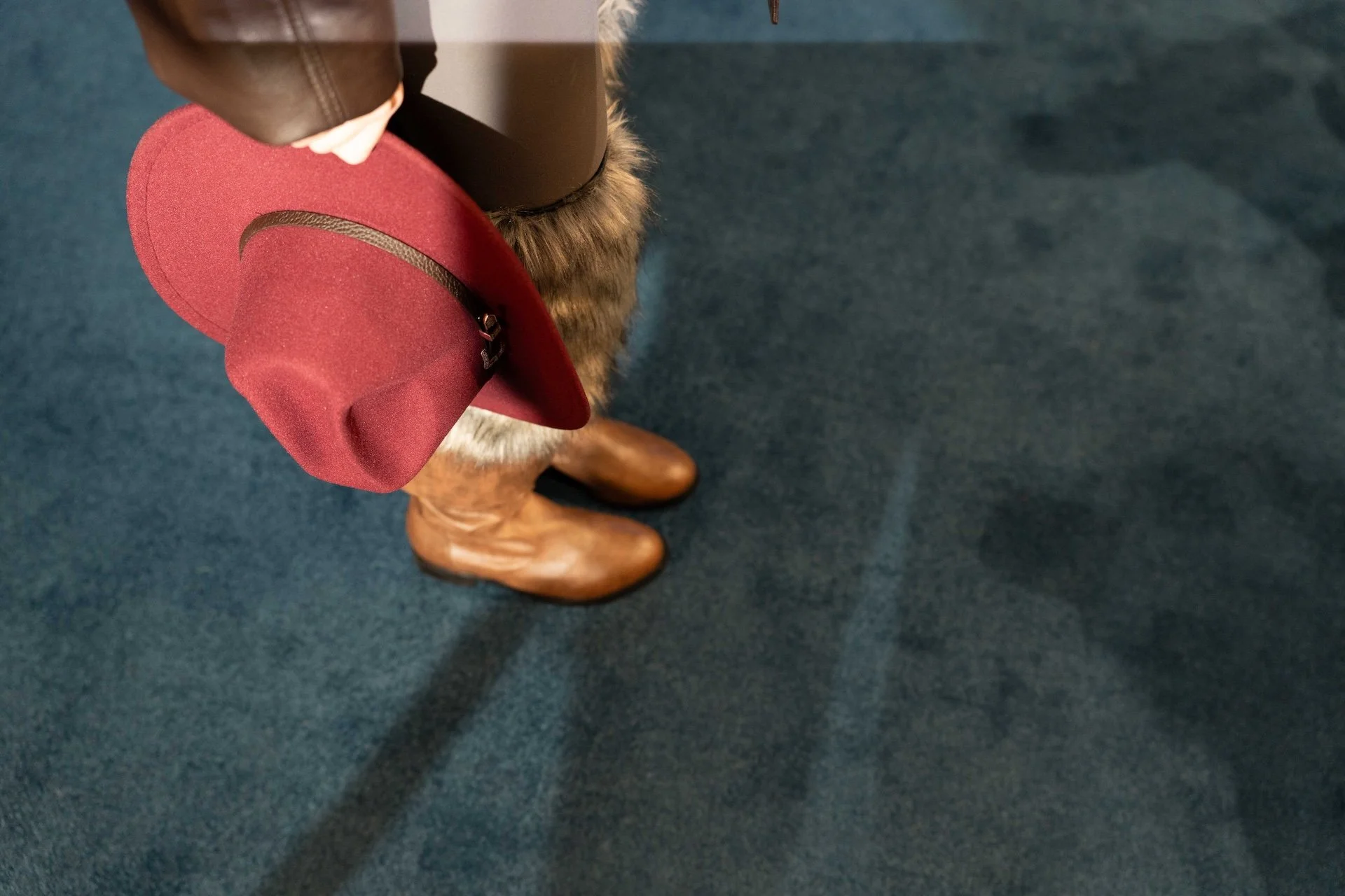 A person standing on a dark blue carpeted floor, wearing light brown cowboy boots, dark brown pants, a brown jacket, and holding a red cowboy hat with a black band. The person's face is not visible.