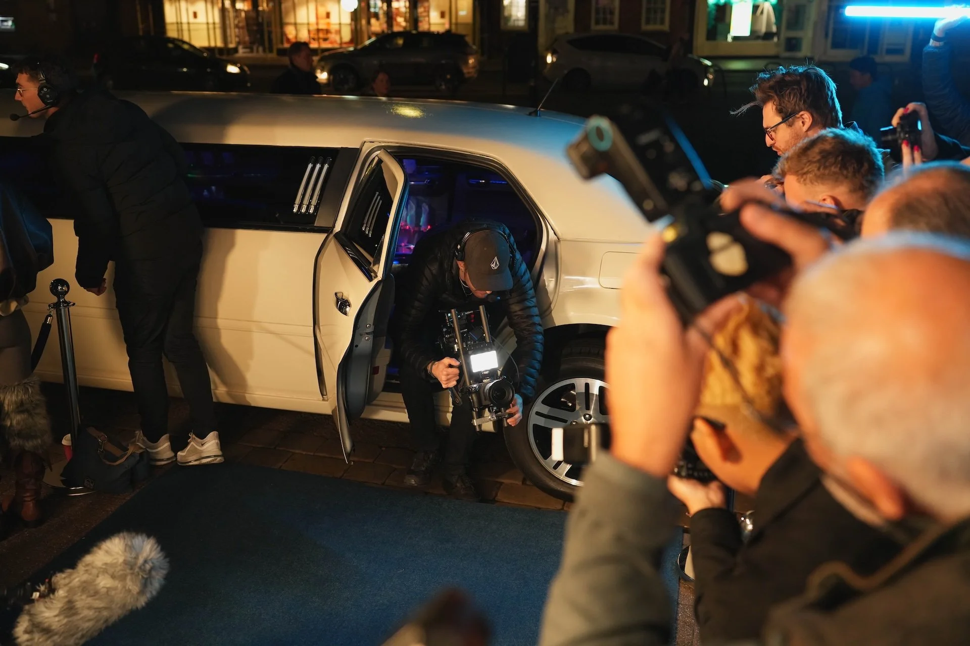 Night scene showing a group of people, including a photographer with equipment, gathered around an open car door in an urban area. City lights and storefronts are visible in the background.