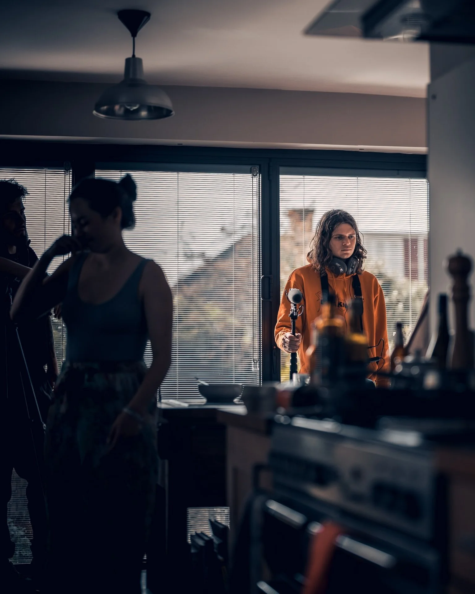 A group of people in a kitchen with large windows and blinds, with one person in an orange hoodie holding a microphone.