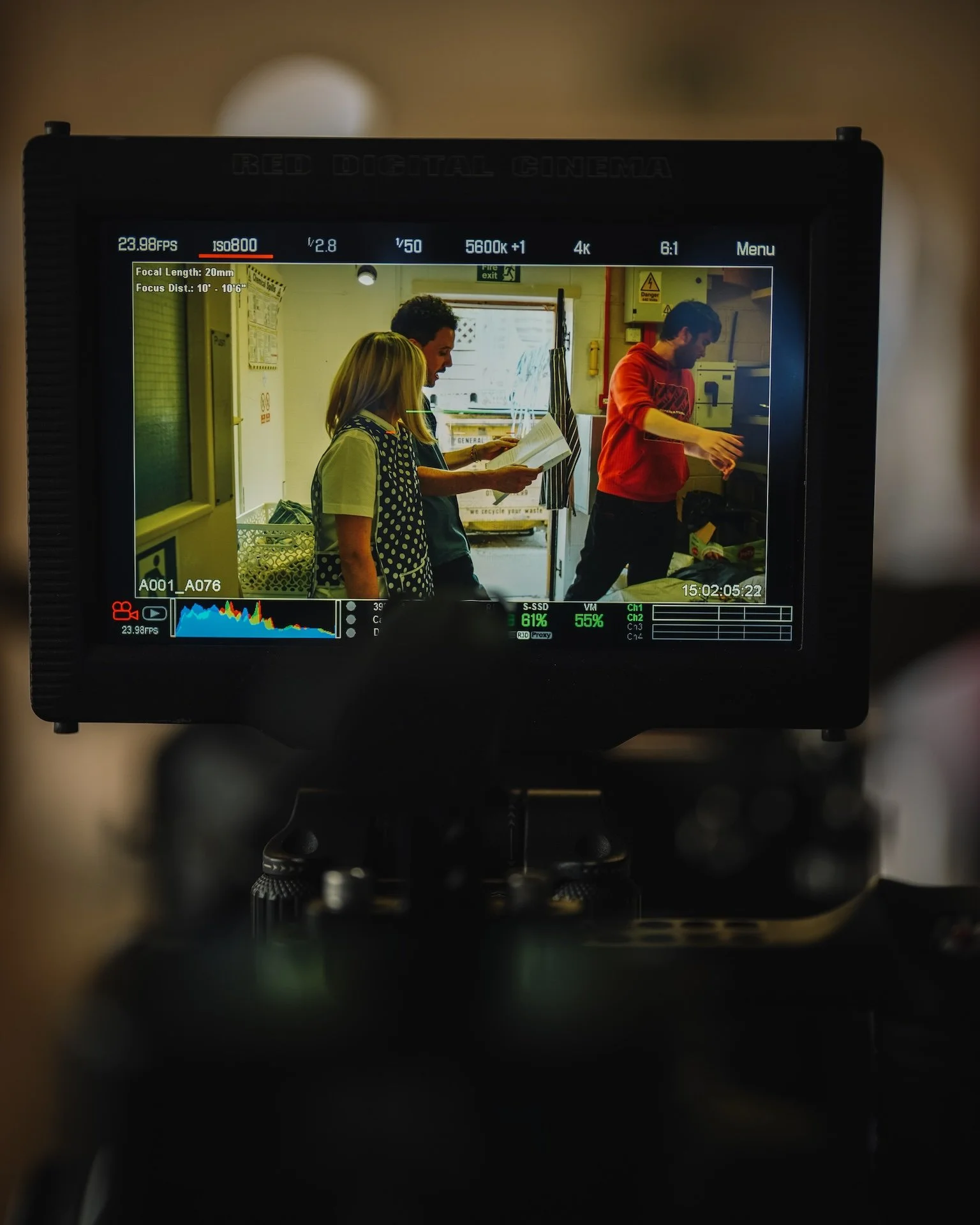 The image shows a camera viewfinder displaying three people inside a room, one woman reading a book, and two men, one in an orange hoodie and the other in a dark shirt, standing near a counter or appliance.