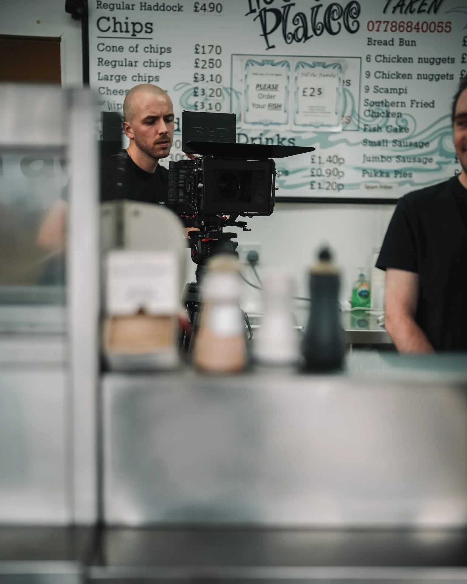 A person with a shaved head working behind a camera in a food shop kitchen. The background shows a menu with prices for chips, fish, and other foods, and a whiteboard with handwritten prices.