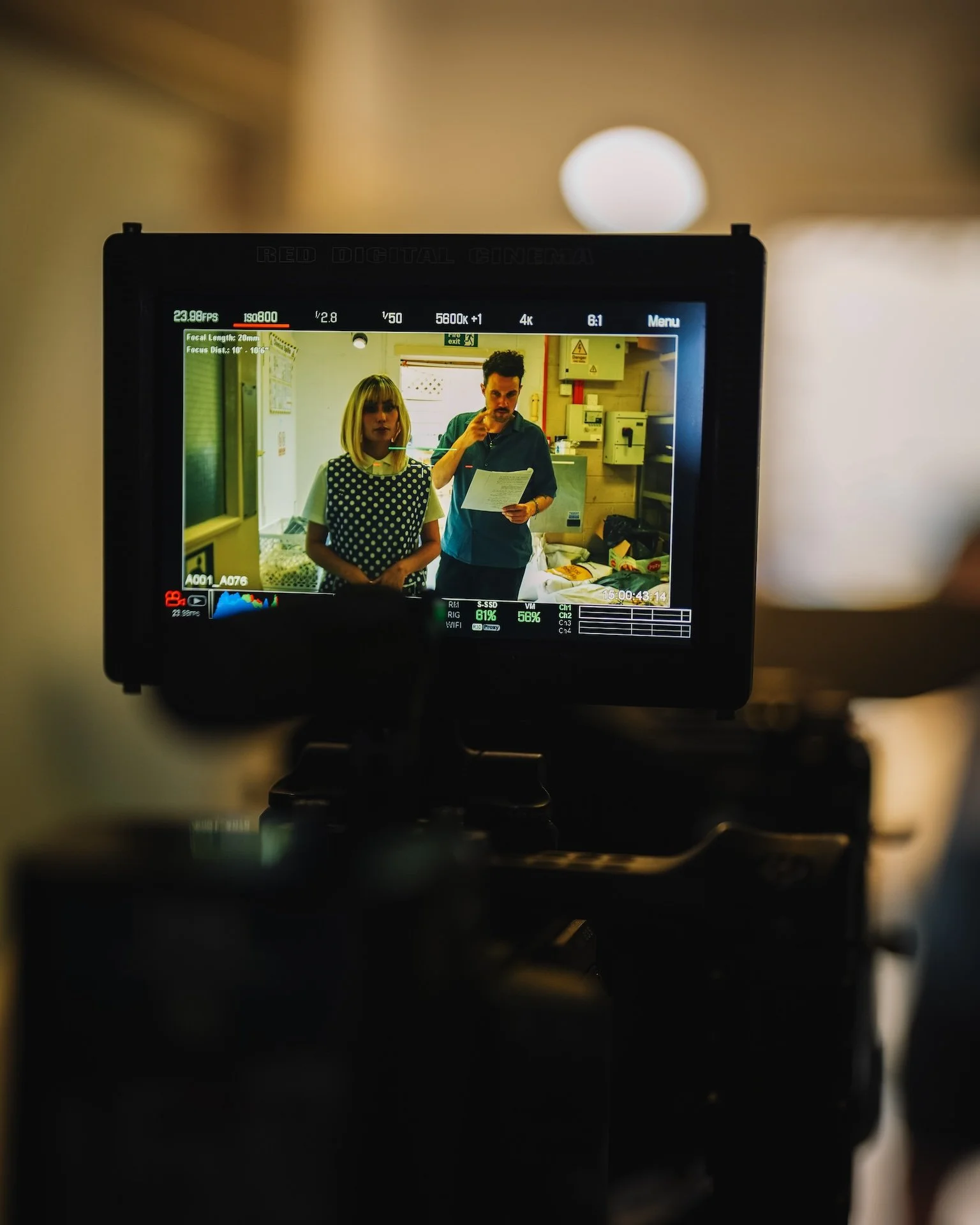 On a camera screen, two people are indoors, a woman with blonde hair and a polka-dot dress and a man with dark hair and a mustache, holding a paper, in a room with electrical equipment, shelves, and boxes.