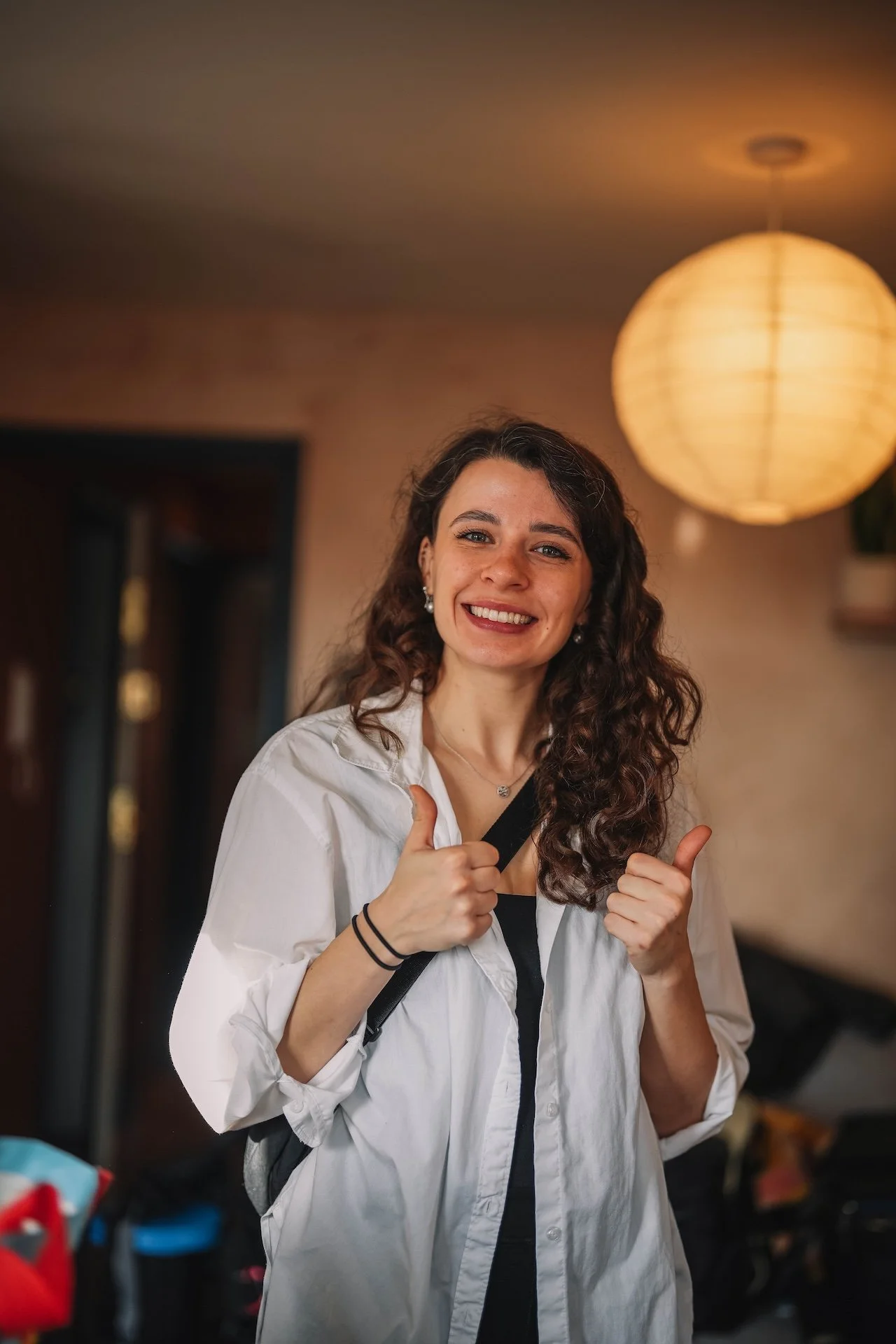 Smiling woman with long, curly brown hair wearing a white shirt and black top, giving two thumbs up indoors with warm lighting.
