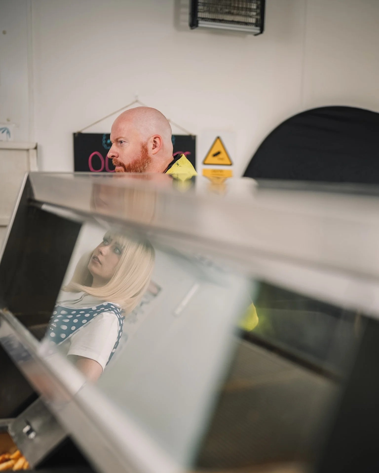 A man with a bald head and red beard looking to the left, and a woman with blonde hair wearing a white top with blue polka dots, reflected in a metallic surface.