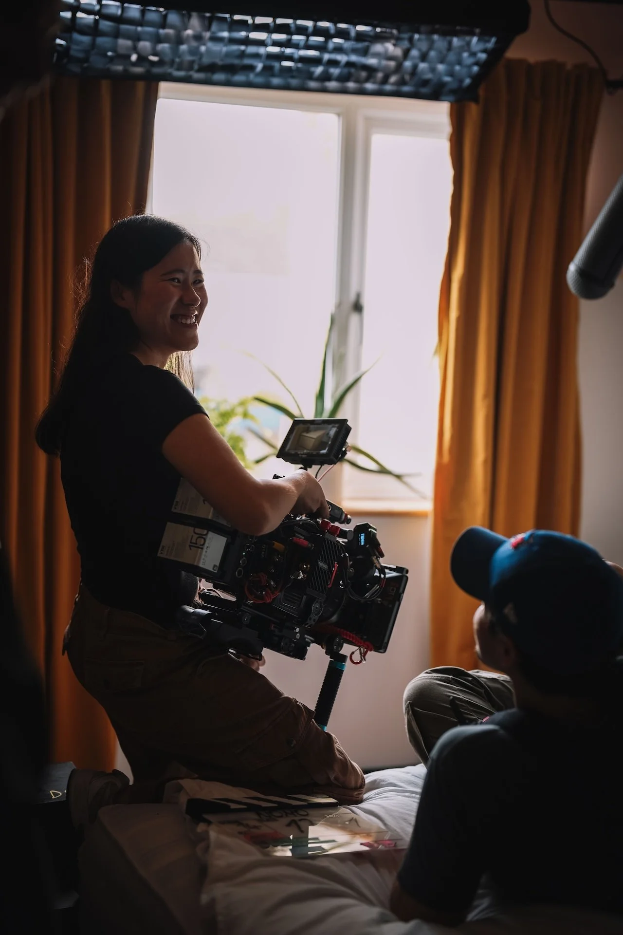 A woman smiling while holding a professional film camera indoors near a window with orange curtains, with two people sitting in front of her.