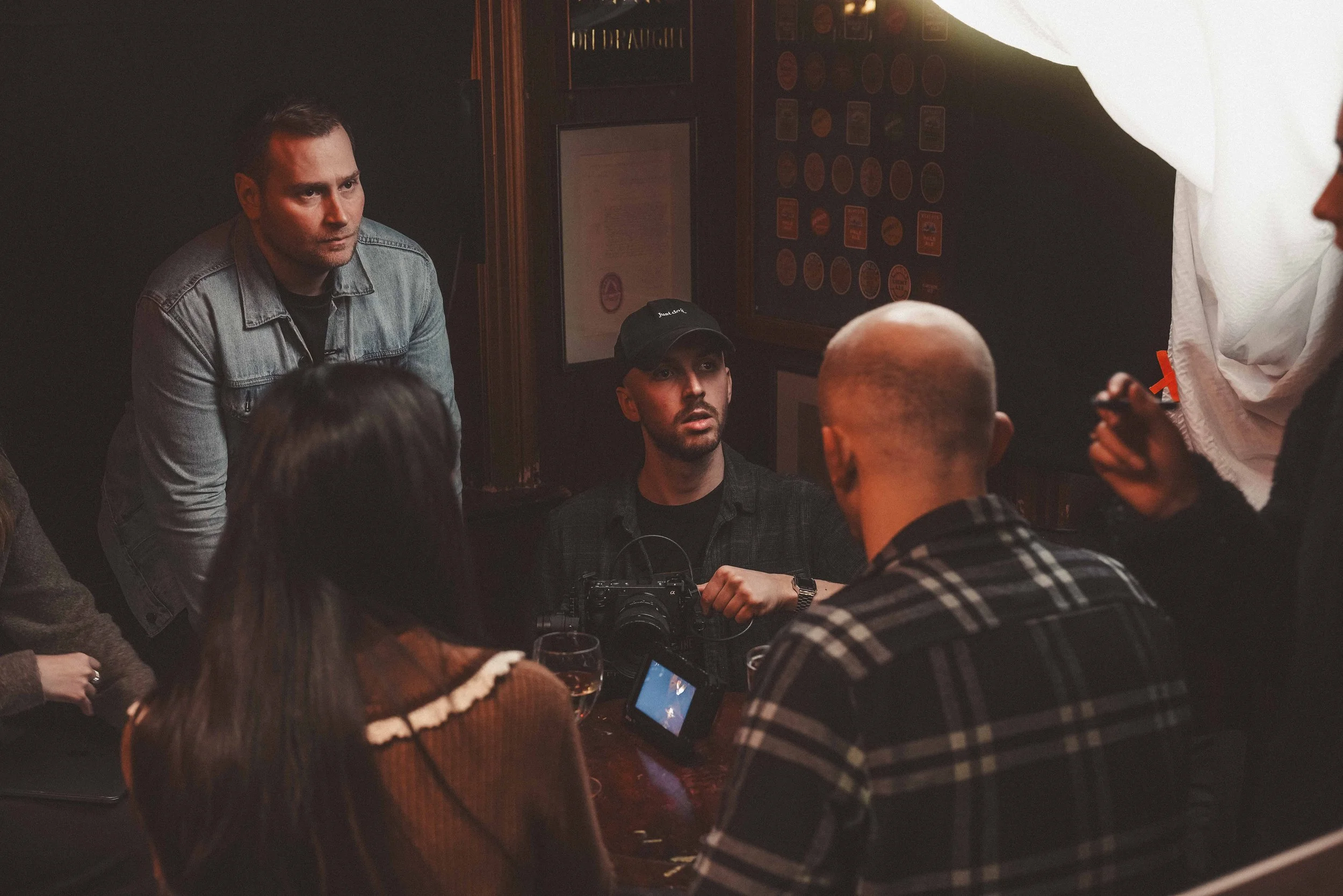 A group of people gathered in a dimly lit bar, with one person standing and others seated, some with drinks and a camera on the table, engaged in conversation.