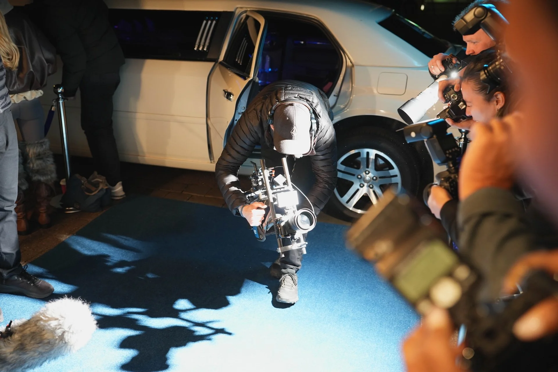 Person operating a camera on a stabilizer in front of a group of photographers taking pictures, outside a white vehicle, at night.