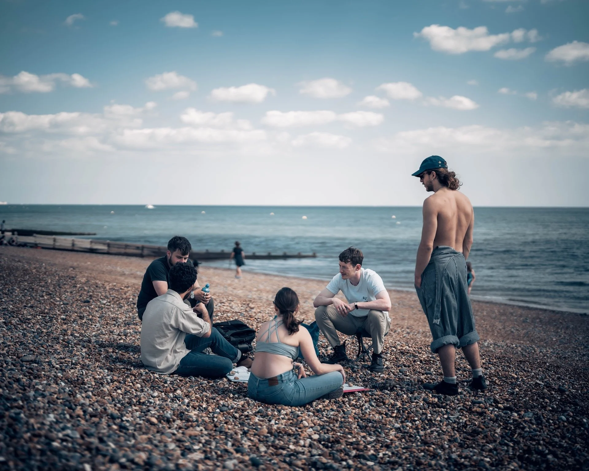 Group of young adults sitting and standing on a pebbled beach near the ocean, with cloudy blue sky overhead, some engaged in conversation and relaxing.