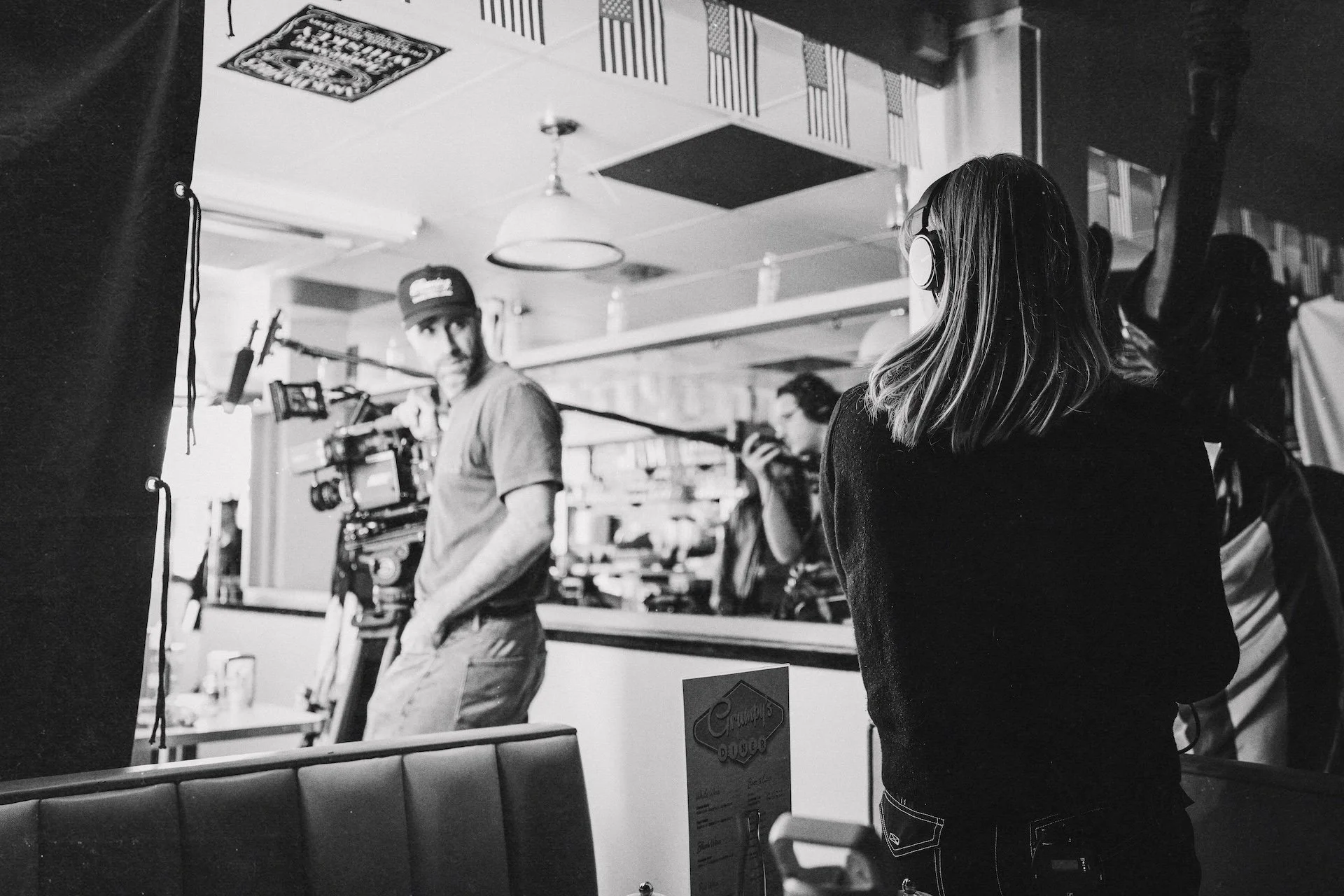 Behind the scenes of a film or television production, a woman with long hair and headphones is observing others preparing to film. A man is holding a camera, and another person is using a walkie-talkie in a well-lit room with a decorated ceiling.