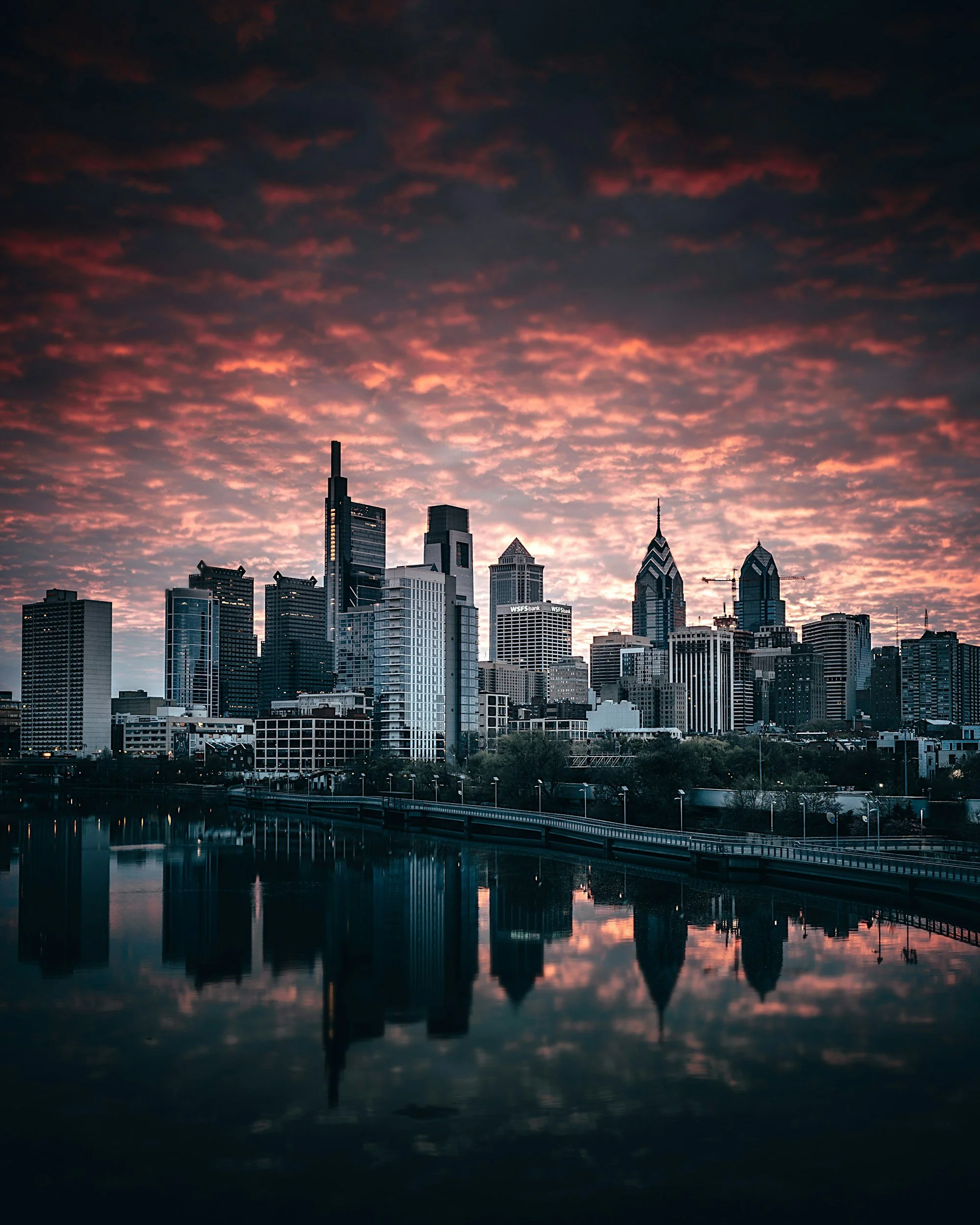 Skyline of a city with tall buildings at sunset or sunrise, with clouds in the sky and their reflection in the water below.