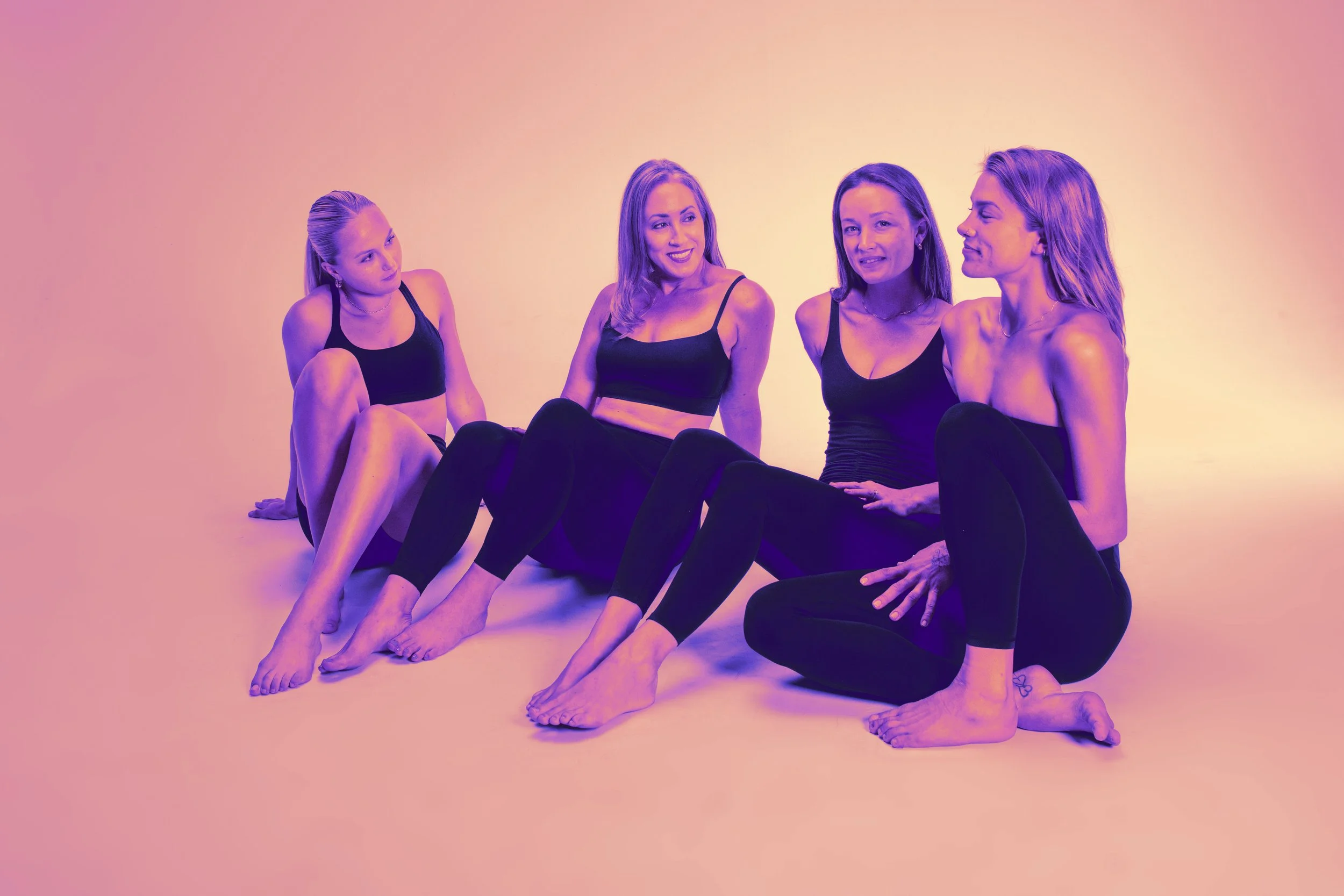 Four women sitting on the floor, engaged in conversation, wearing black athletic wear, against a plain light-colored background with a purple and pink gradient overlay.