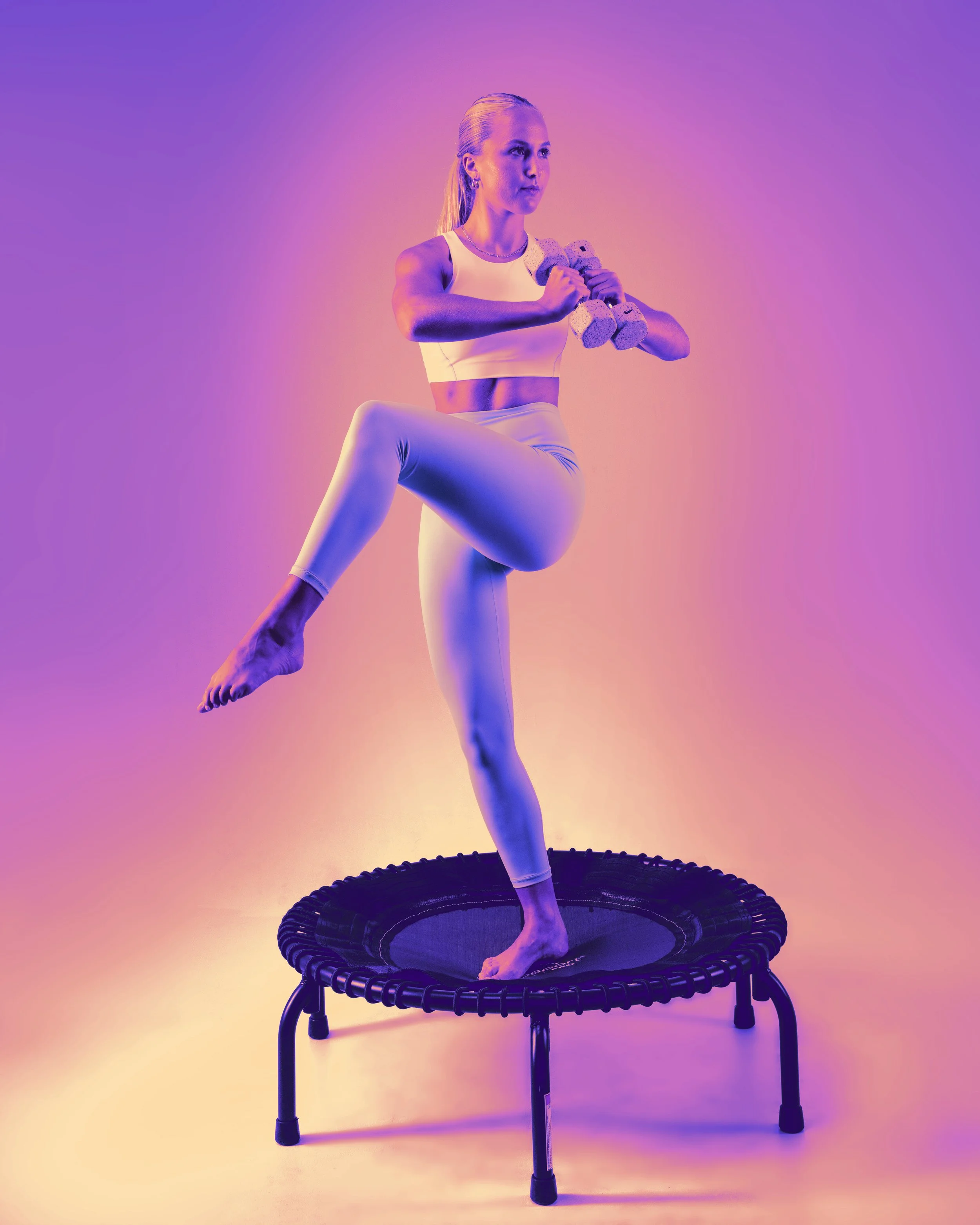 A woman in workout attire balancing on one leg on a mini trampoline while holding dumbbells with pink, purple, and yellow lighting.
