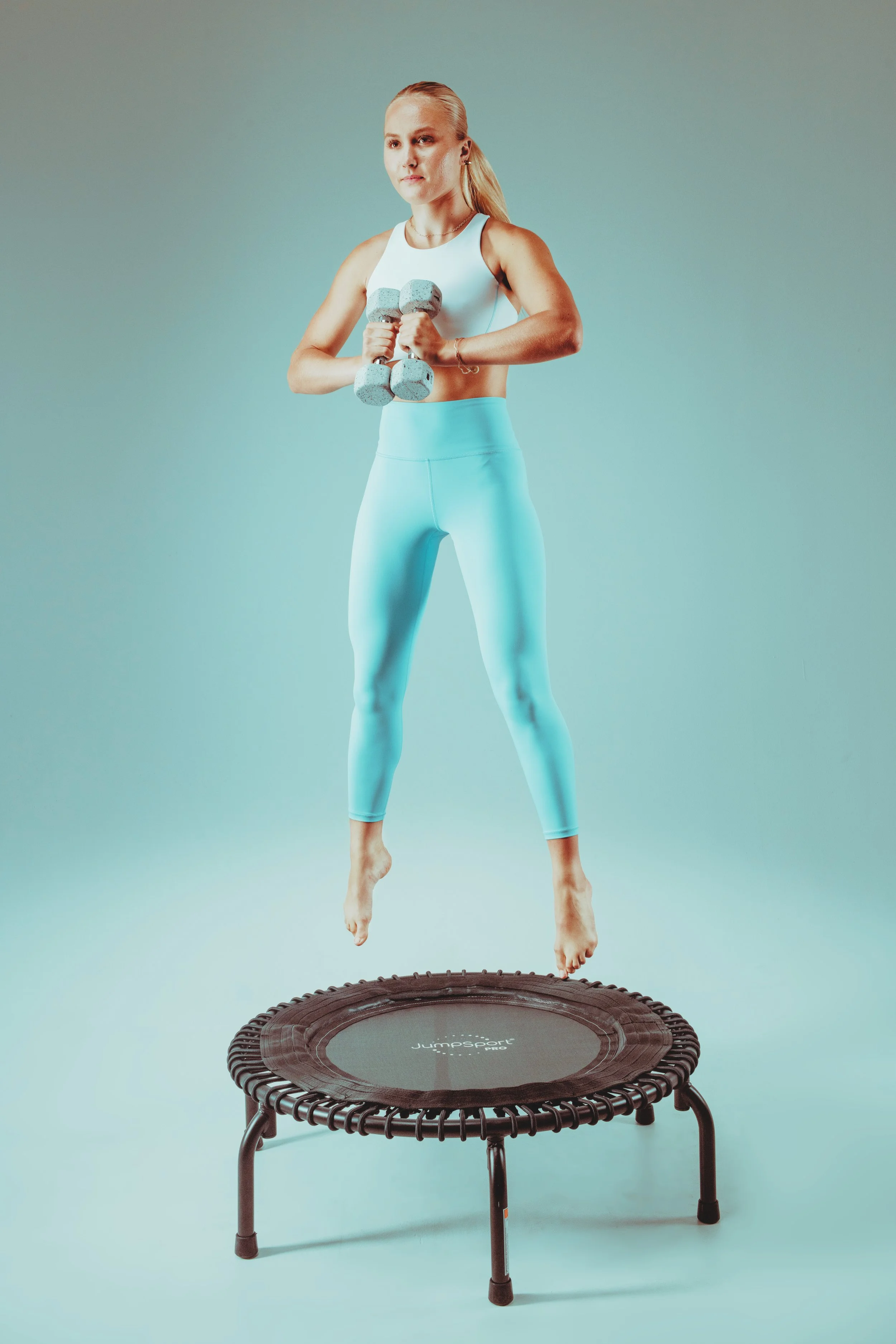 A woman in light blue leggings and a white sports top jumping on a mini trampoline while holding a small set of dumbbells.
