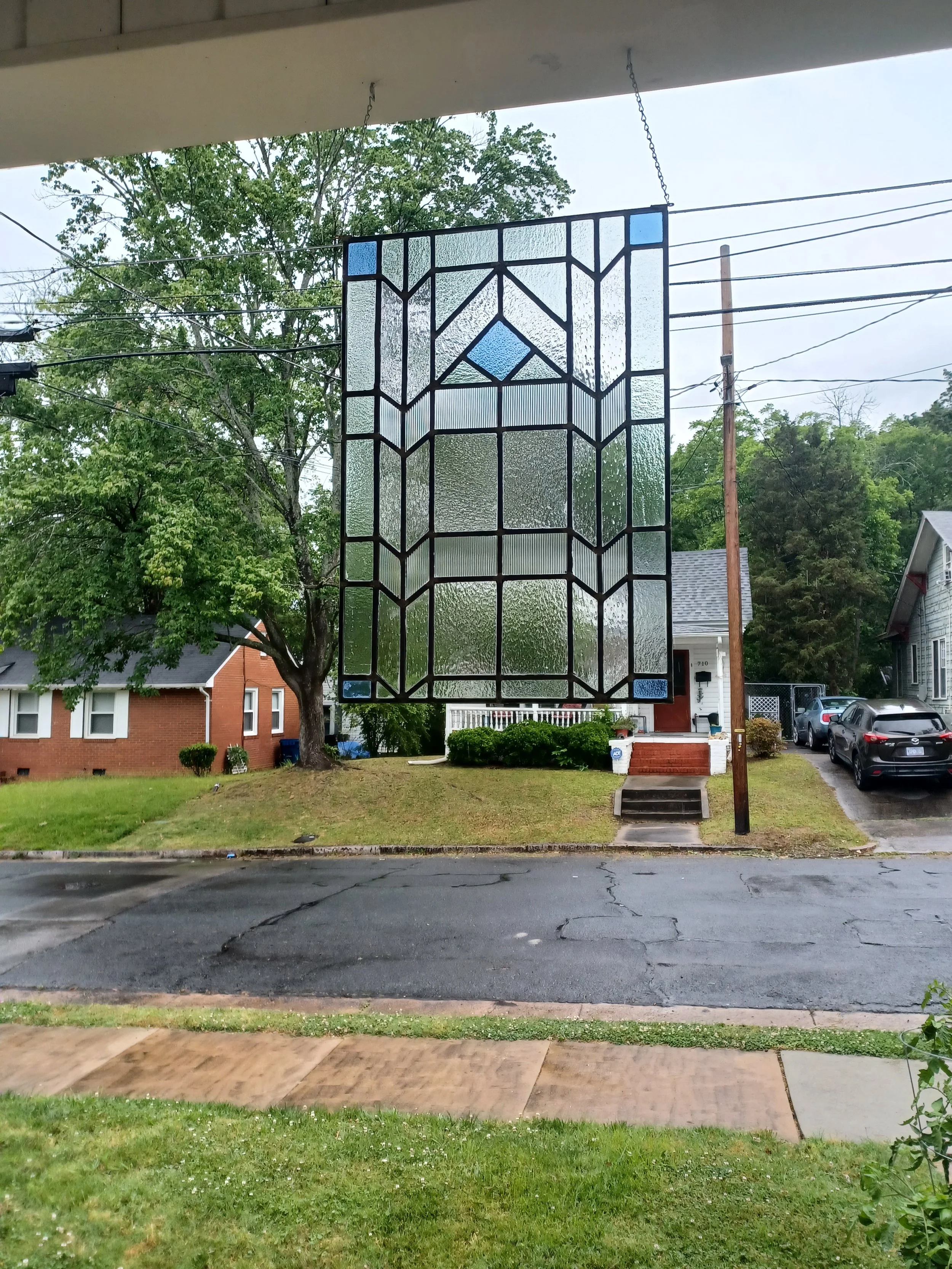 A stained glass panel hanging outside on a porch with houses and trees in the background.