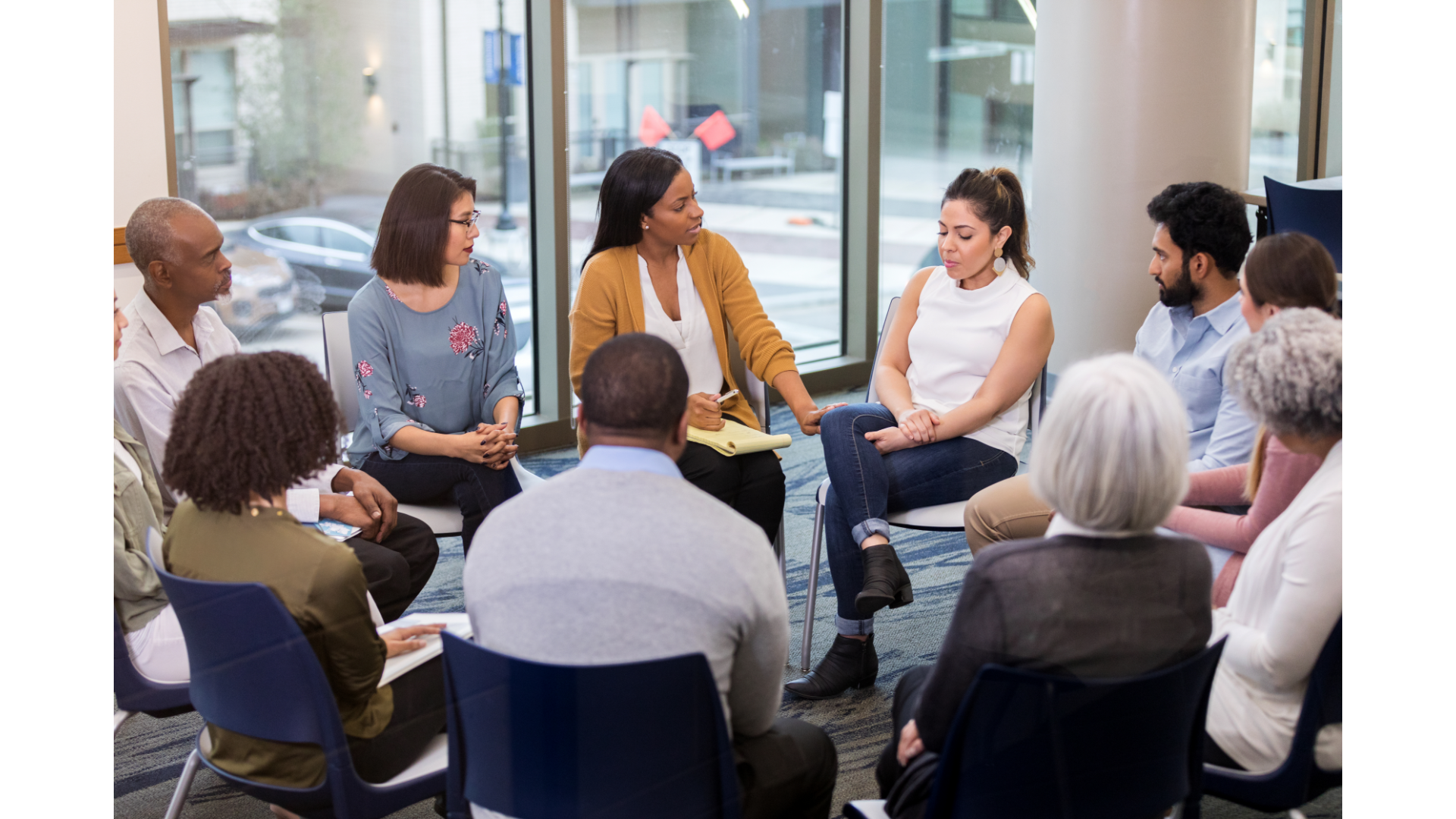 Group of six diverse people sitting in chairs in a circle, engaging in a discussion or support group session in a bright room.