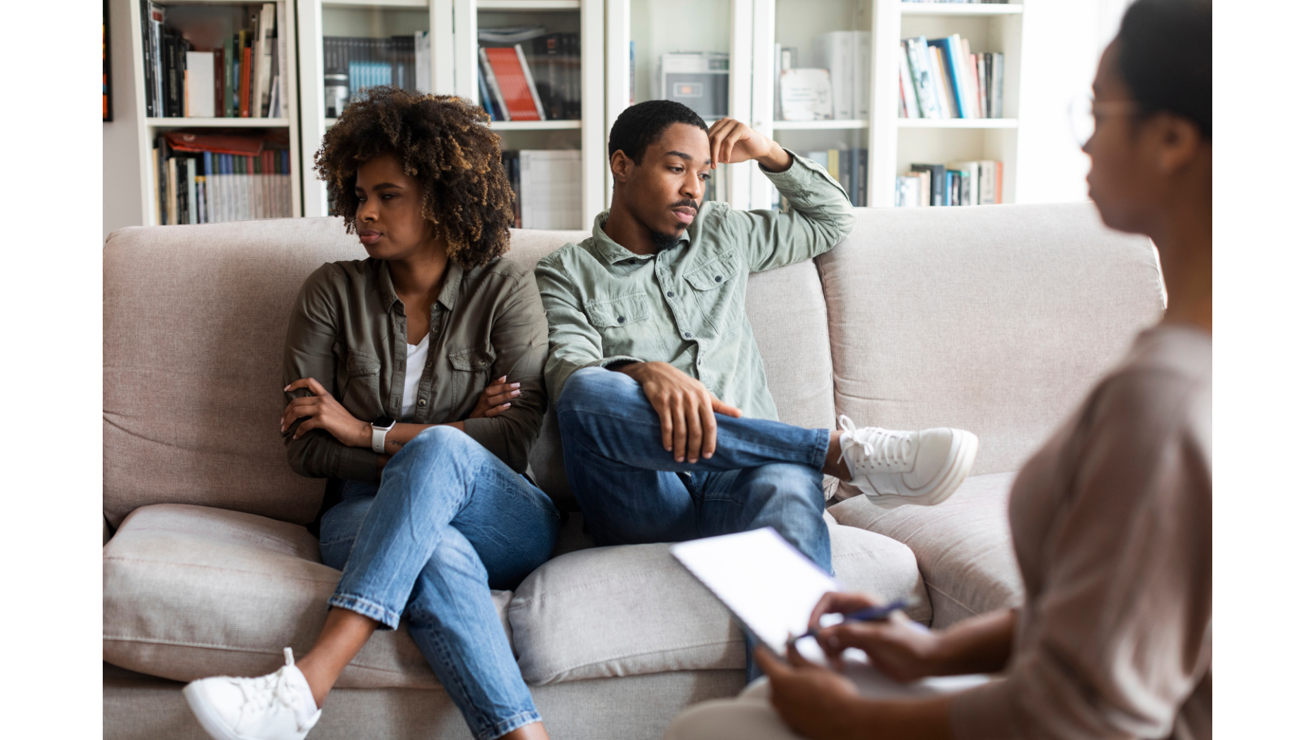 A woman in a cream sweater and jeans sits with arms crossed, looking distressed during a counseling session. A female therapist with a notepad listens attentively, and a male partner in a denim shirt and beige pants sits on a beige sofa with hands clasped.