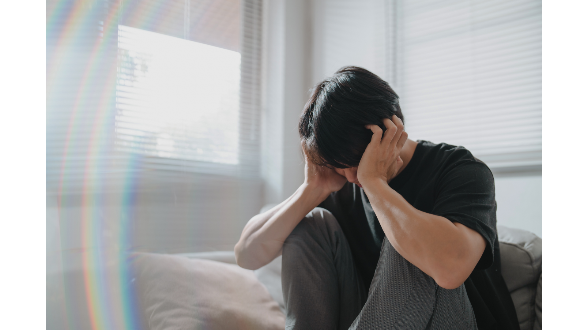 A person sits on a couch with their head in their hands, leaning forward in a moment of stress or overwhelm. The scene reflects emotional exhaustion, anxiety, and the quiet weight many people carry alone.