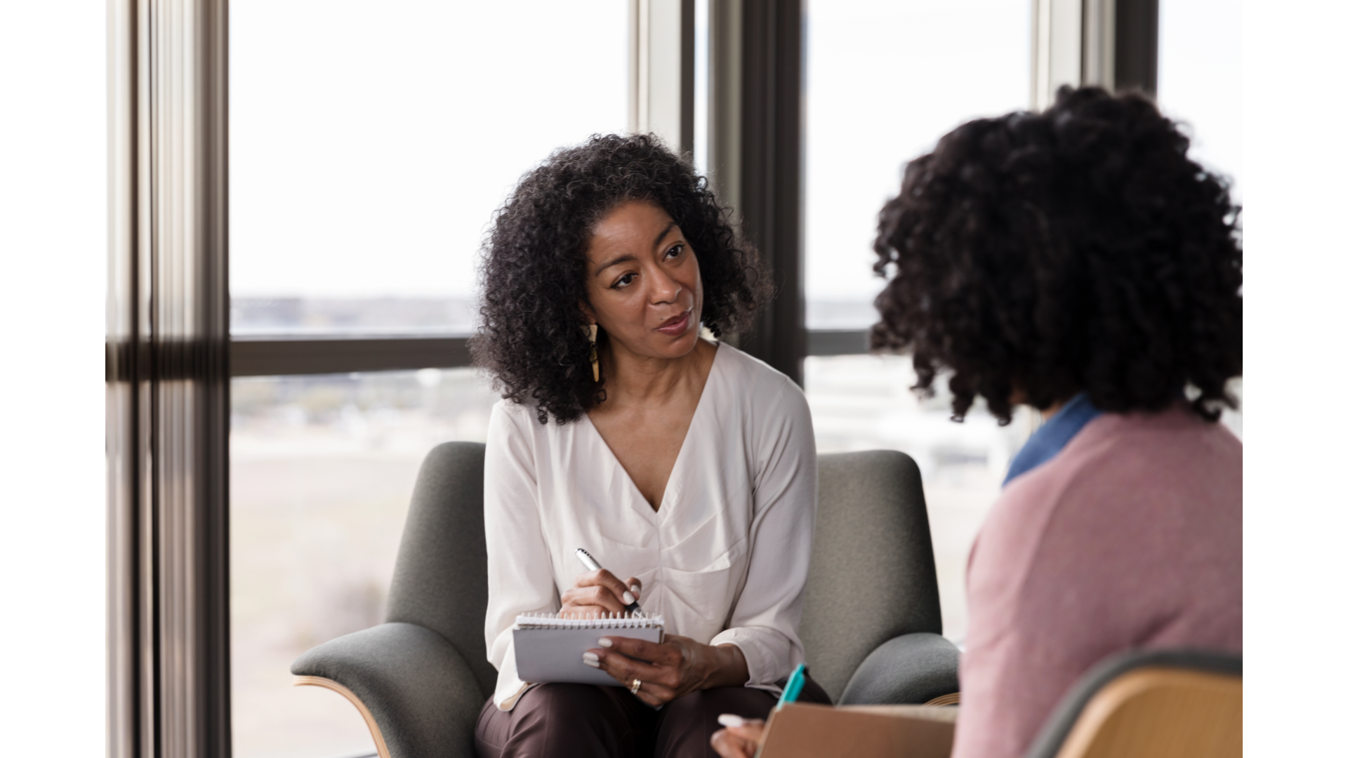 A woman in a beige long-sleeve shirt appears distressed during a therapy session with a therapist, who is taking notes on a notepad.