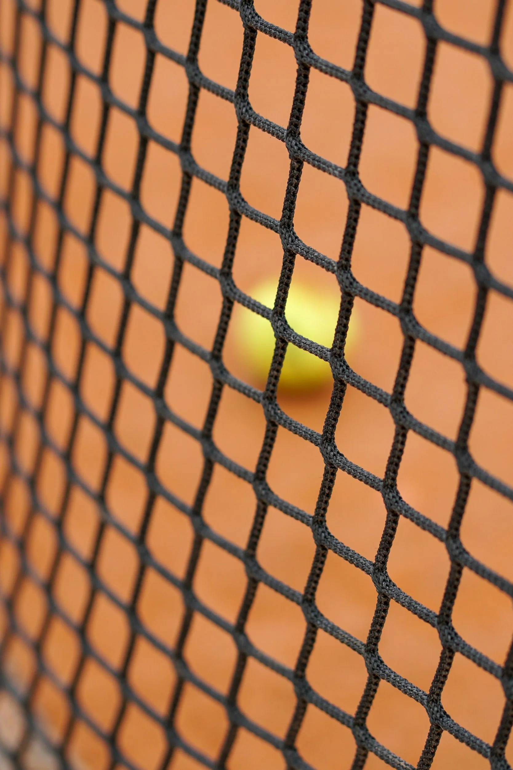 Nahaufnahme eines schwarzen Tennisnetzes mit einem gelben Tennisball, der durch das Netz sichtbar ist, auf einem Tennisplatz.