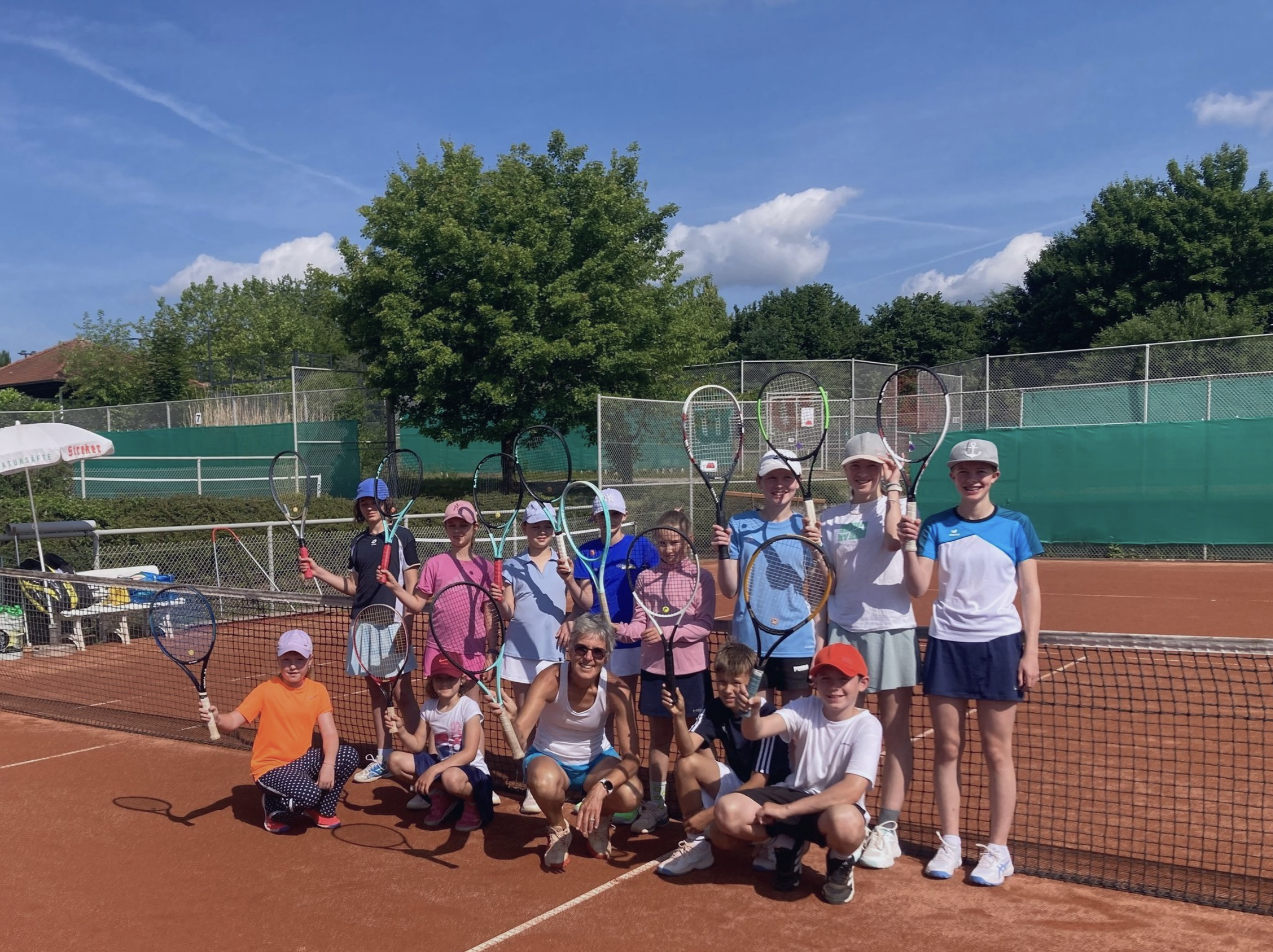 Gruppe von Kindern und einer erwachsenen Person auf einem Tennisplatz, alle mit Tennisracket, Sonne und blauer Himmel.
