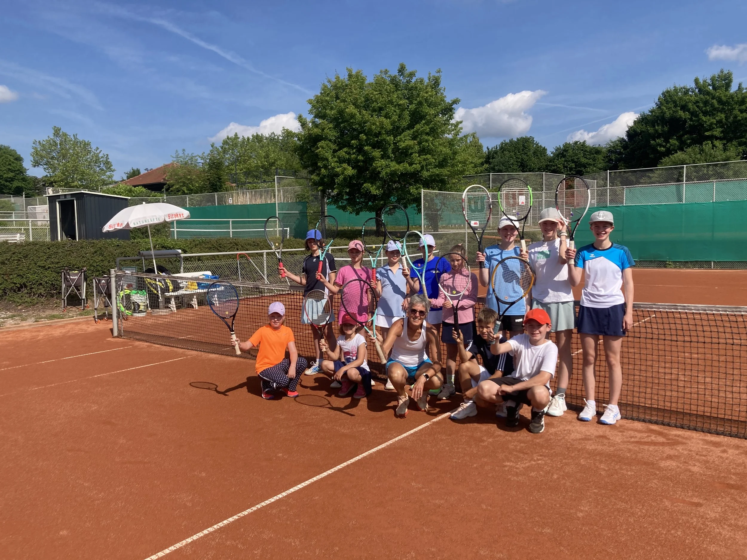Eine Gruppe von Kindern und einer Frau auf einem Tennisplatz, alle halten Tennisschläger und tragen Sportkleidung und Hüte, bei sonnigem Himmel.