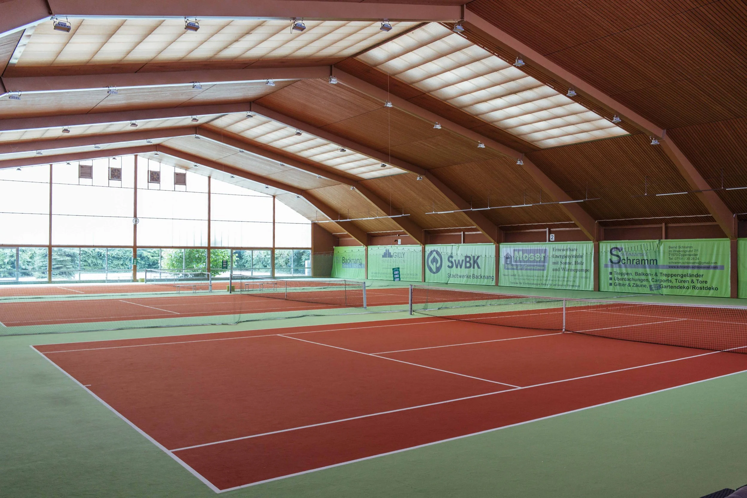 Innenansicht eines modernen Tennisplatzes mit roten und grünen Belägen, großen Fenstern und Werbebannern an der Wand