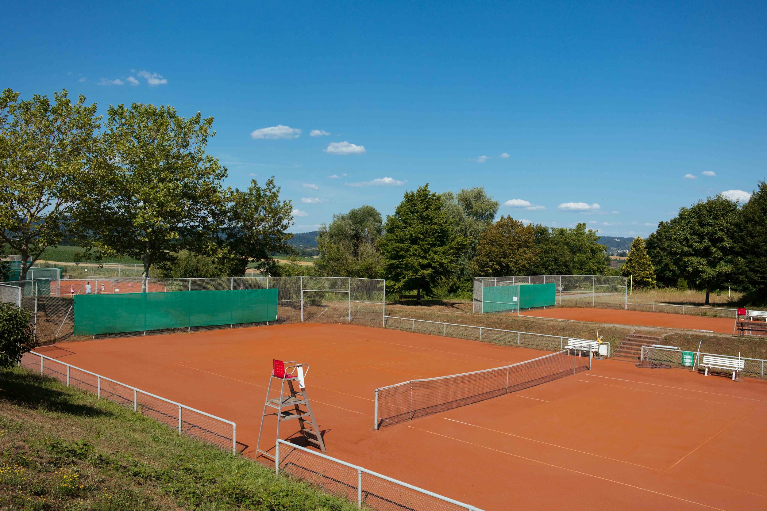 Zwei Tennisplätze mit roter Oberfläche, umgeben von Bäumen und einem blauen Himmel.