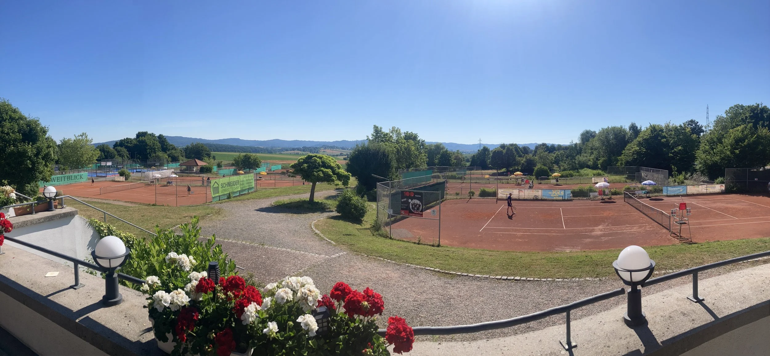 Tennisplätze im Freien auf einem sonnigen Tag, mit Spielern und Sonnenschirmen, umgeben von grünen Bäumen und Bergen im Hintergrund.