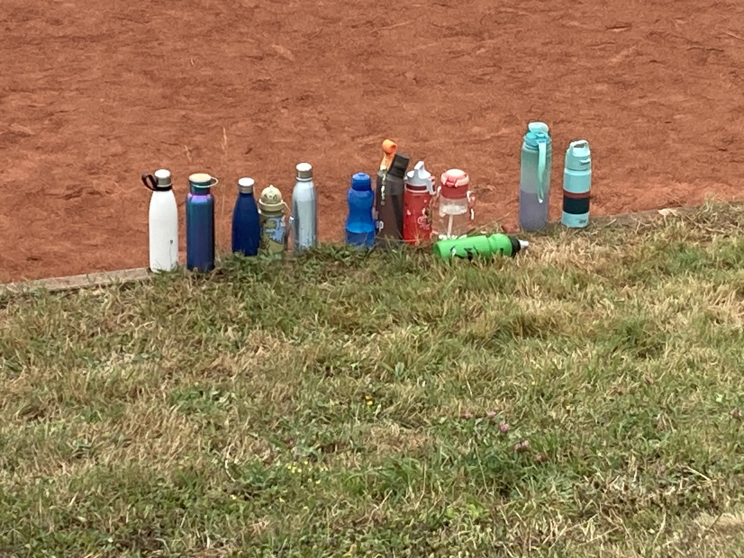 Mehrere Trinkflaschen in unterschiedlichen Farben nebeneinander auf einer Mauer, im Vordergrund Gras, im Hintergrund eine rote Clay-Court.