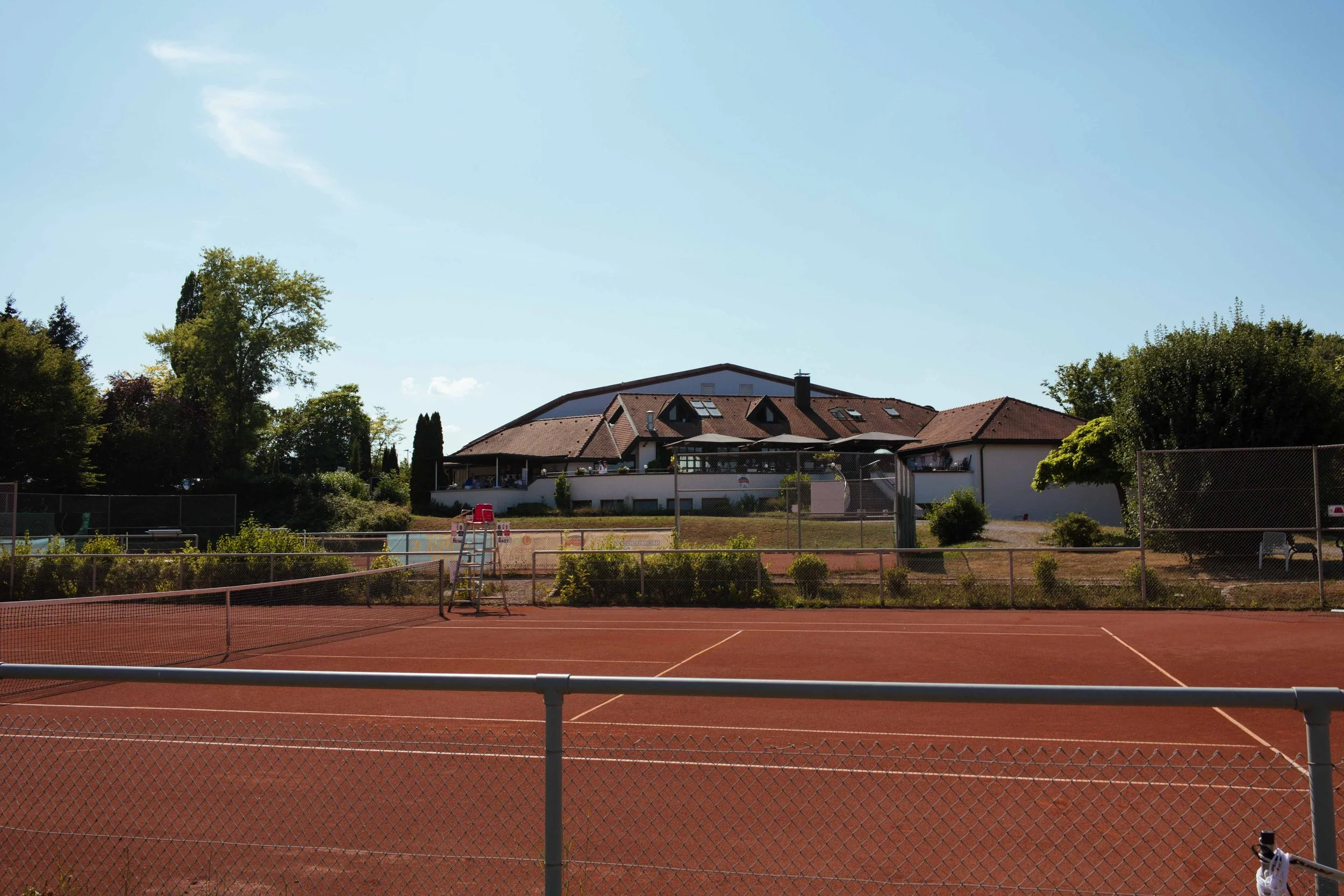 Leerer Tennisplatz vor einem Haus mit Terrasse, umgeben von Bäumen bei sonnigem Wetter.