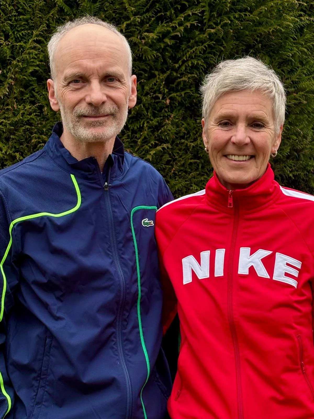 Ein Mann und eine Frau tragen Sportjacken stehen vor einer grünen Hecke