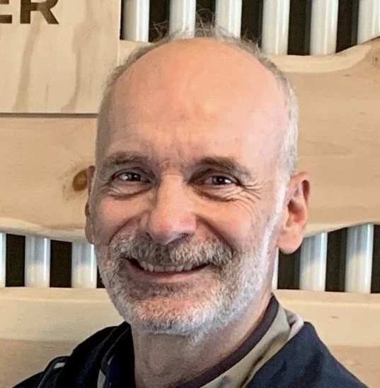 Lächelnder Mann mit grauem Bart und kurzen Haaren vor einer Holzwand.