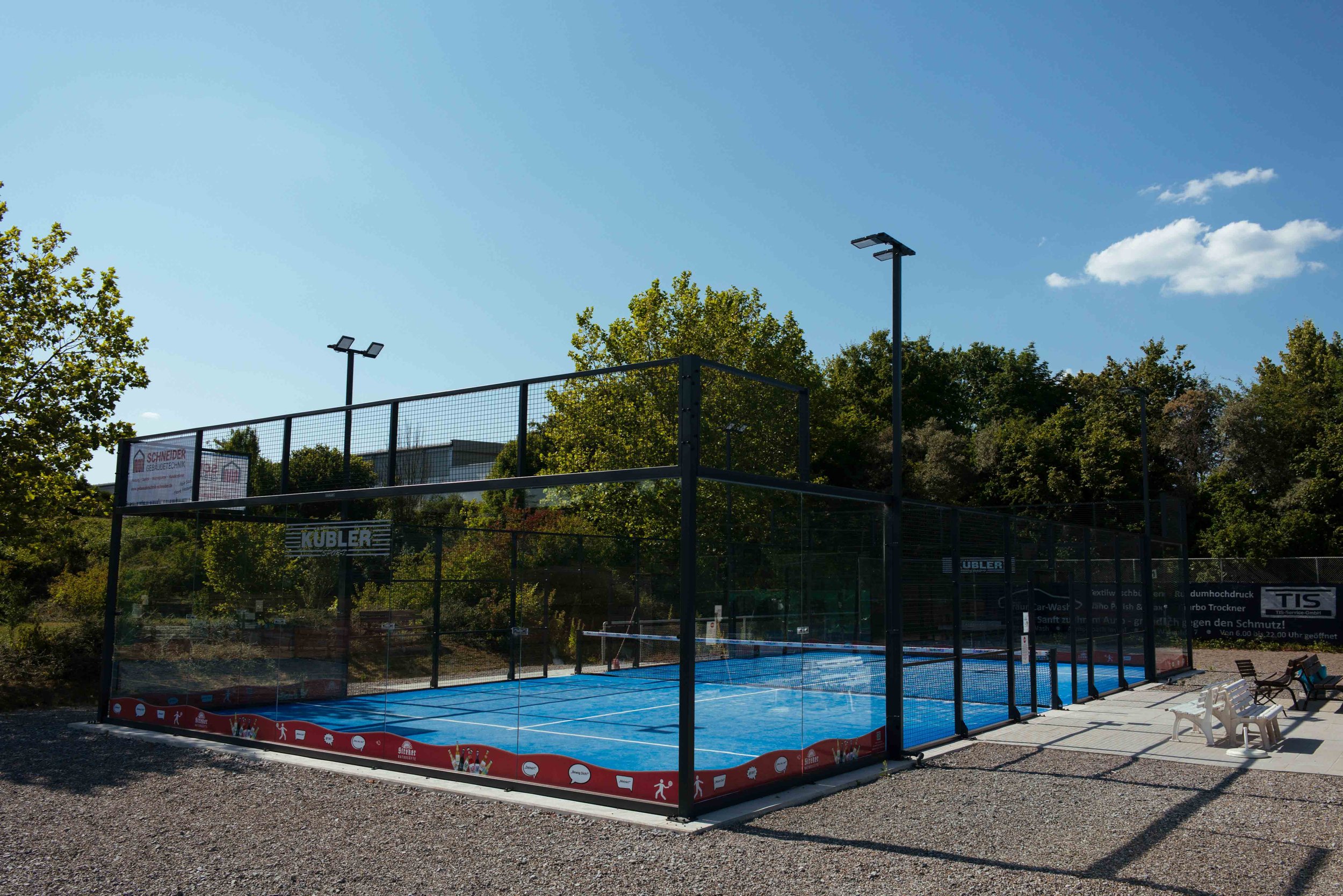 Außen-Tennishartplatz mit blauer Oberfläche und umlaufender Begrenzung, umgeben von Bänken, im Hintergrund Bäume und ein blauer Himmel.