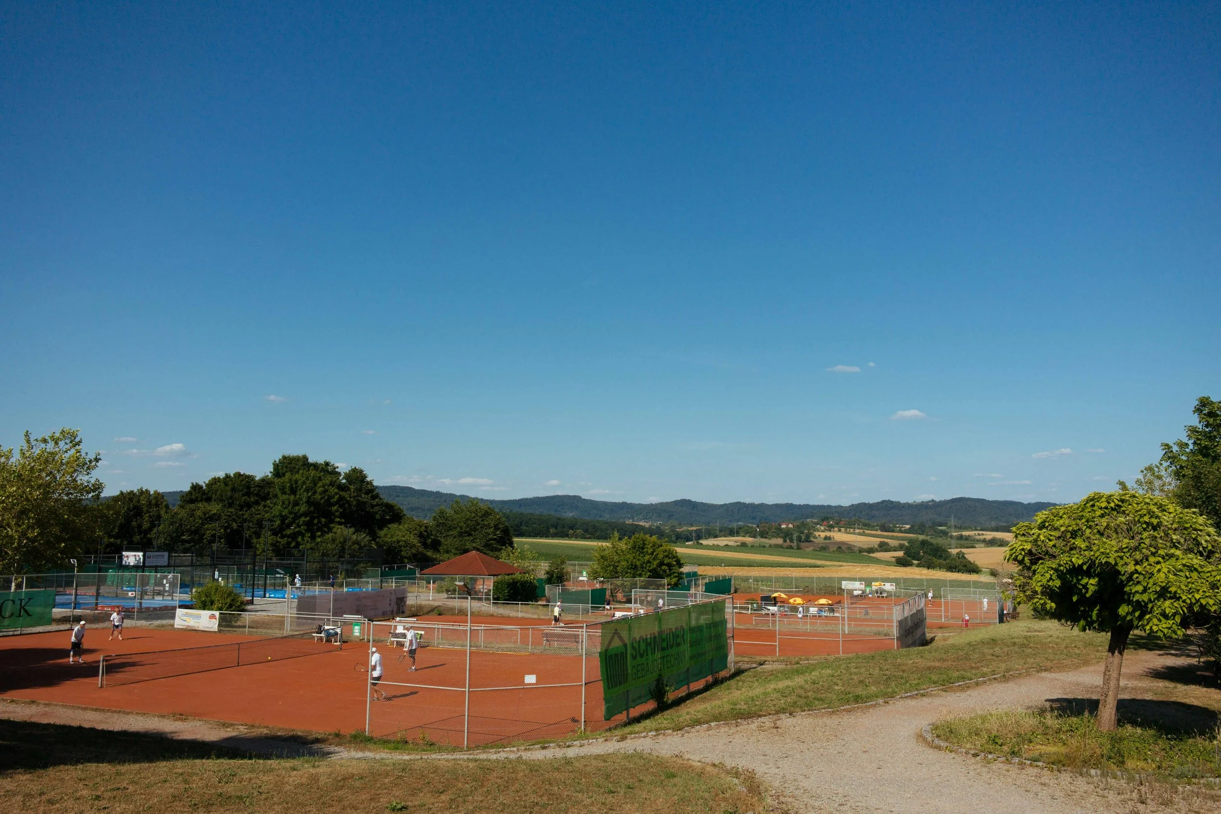 Tennisplätze im Freien mit Spielern, umgeben von grünen Bäumen und Hügeln bei schönem Wetter.