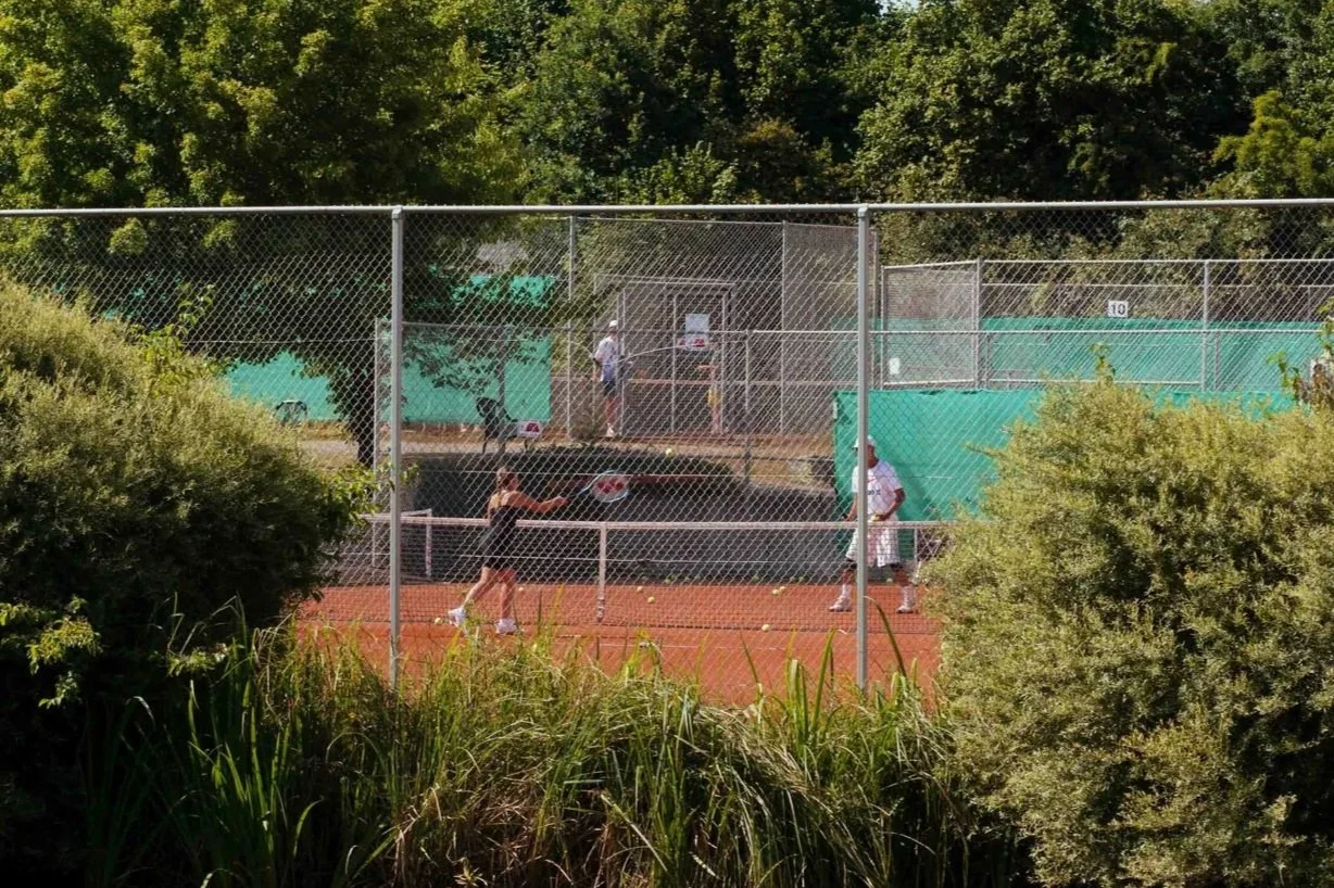 Eine Tennisanlage mit einem Tennisplatz, einige Leute spielen Tennis, im Hintergrund sind Bäume und grüne Wände.