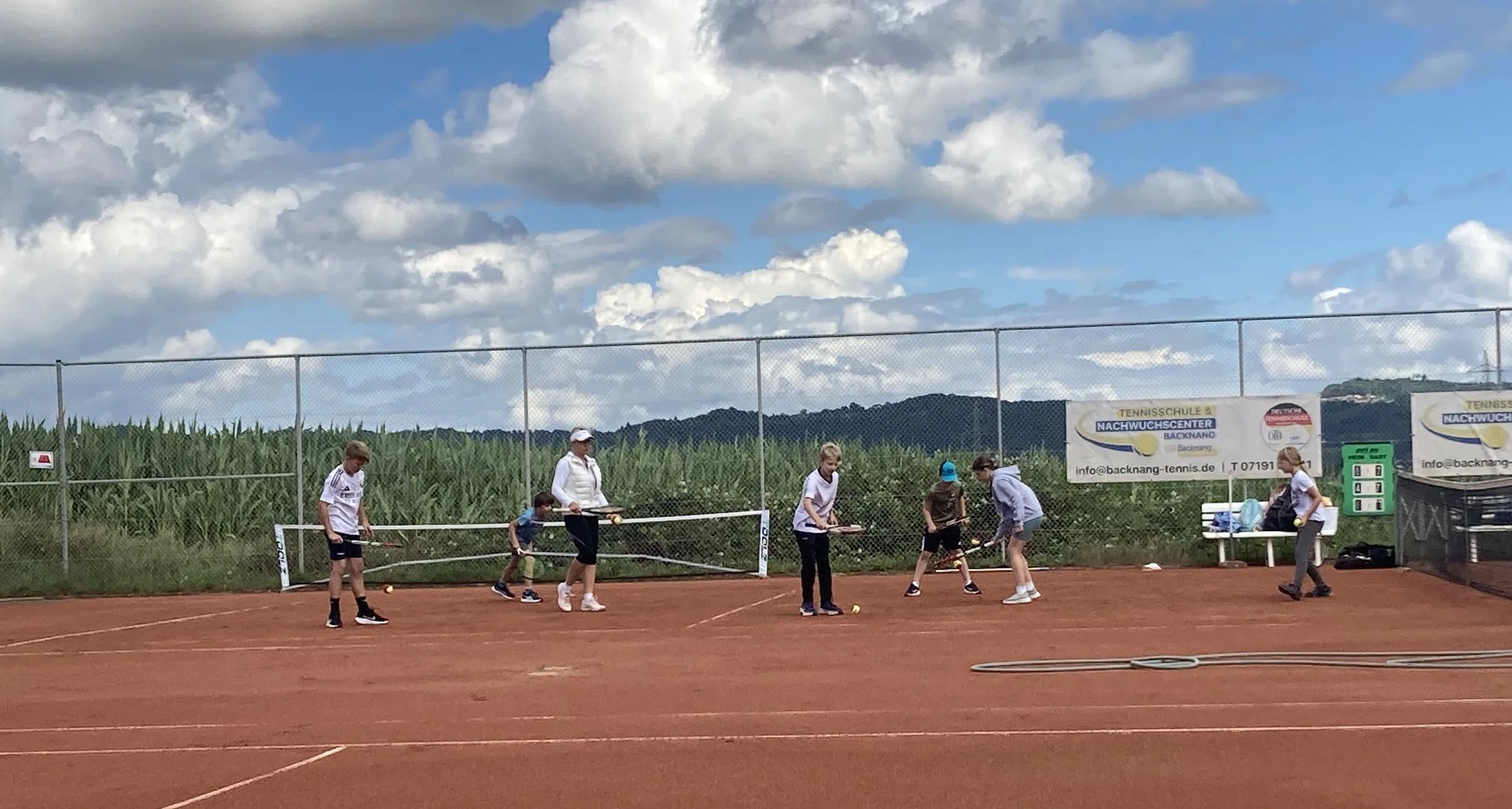 Kinder und Erwachsene beim Tennis auf einem roten Tennisplatz im Freien, mit einem Himmel voller Wolken im Hintergrund.