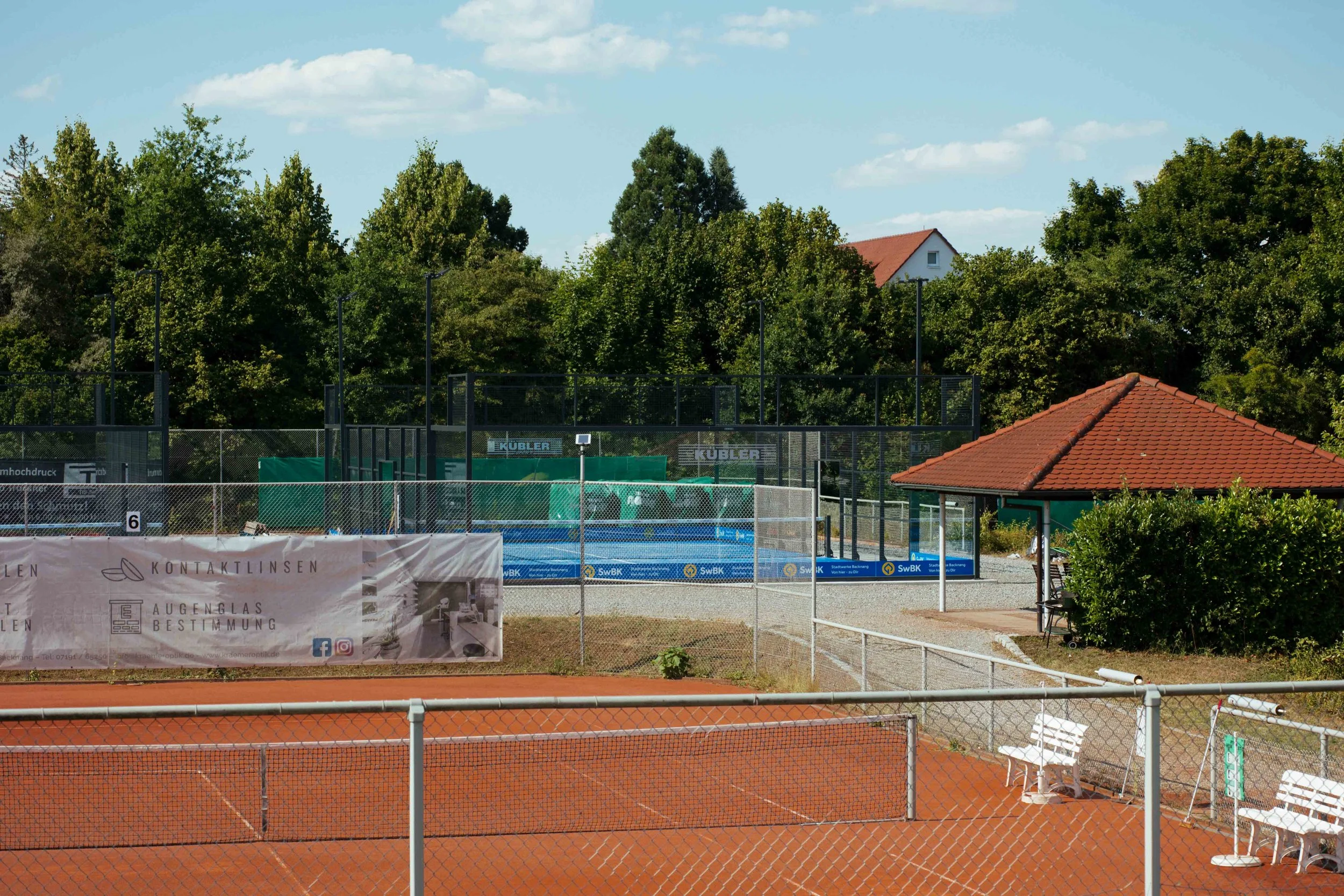 Ein Tennisplatz mit roten Sandboden, umzäunt von einem Drahtzaun, im Hintergrund grüne Bäume und ein kleiner Gebäudekomplex mit rotgefliesten Dach.