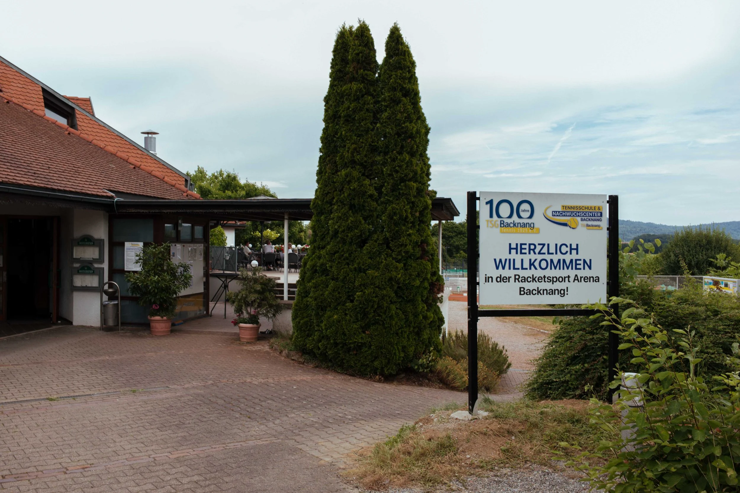 Eingang zur Tennis- und Nachwuchssportschule Backnang mit Begrüßungsschilder. Im Hintergrund ein Tennisplatz mit Spielern und Sitzgelegenheiten.