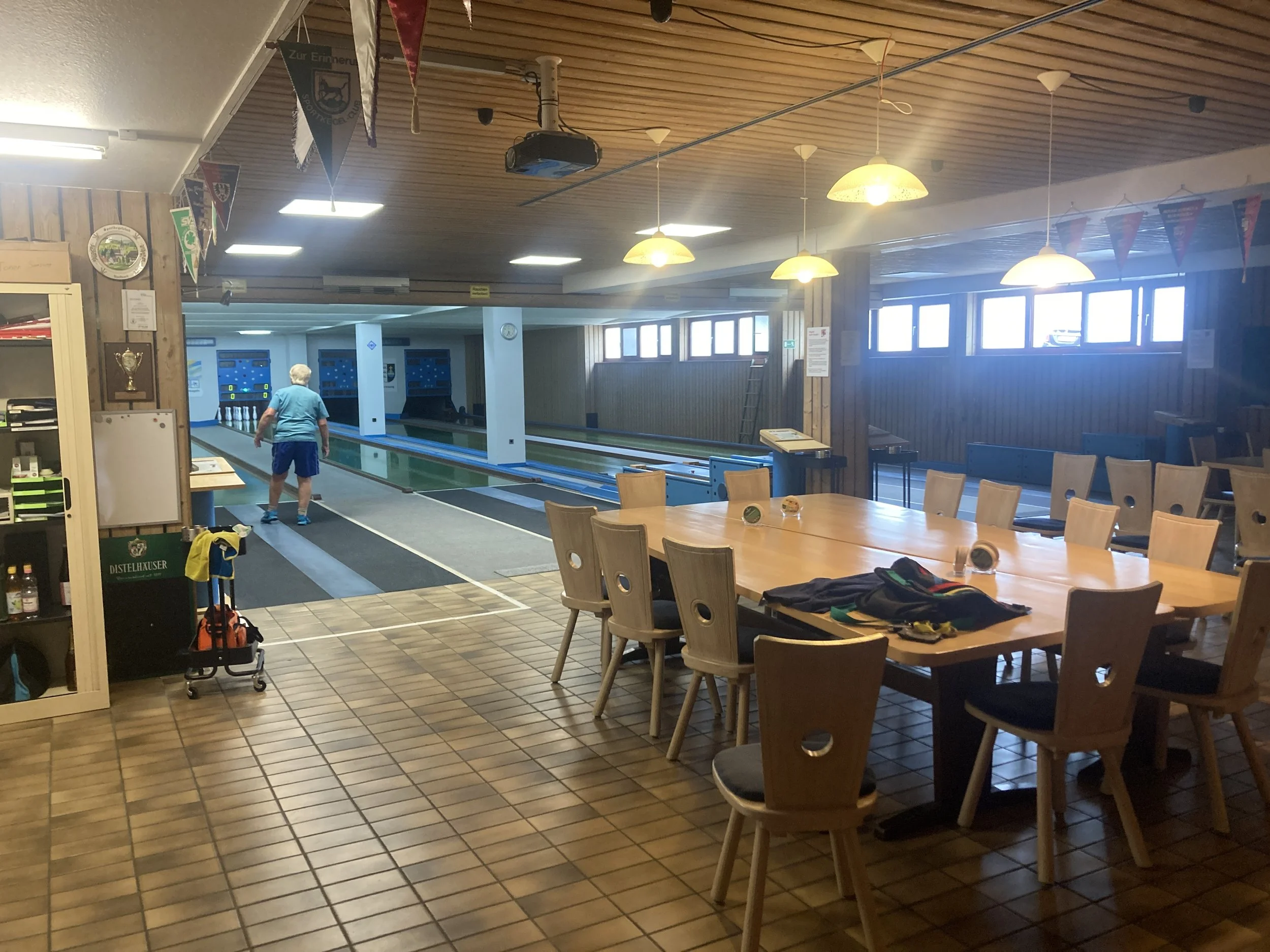 Innenansicht eines Bowlingclubs mit mehreren Bowlingbahnen, einem Mann beim Bowlen, einem großen Tisch mit Stühlen und einigen Gegenständen darauf.