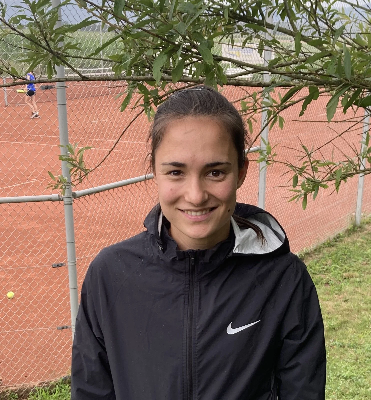 Junge Frau steht auf einem roten Tennisplatz bei sonnigem Himmel, Tennisbälle liegen auf dem Boden, im Hintergrund zwei weitere Personen auf dem Platz.