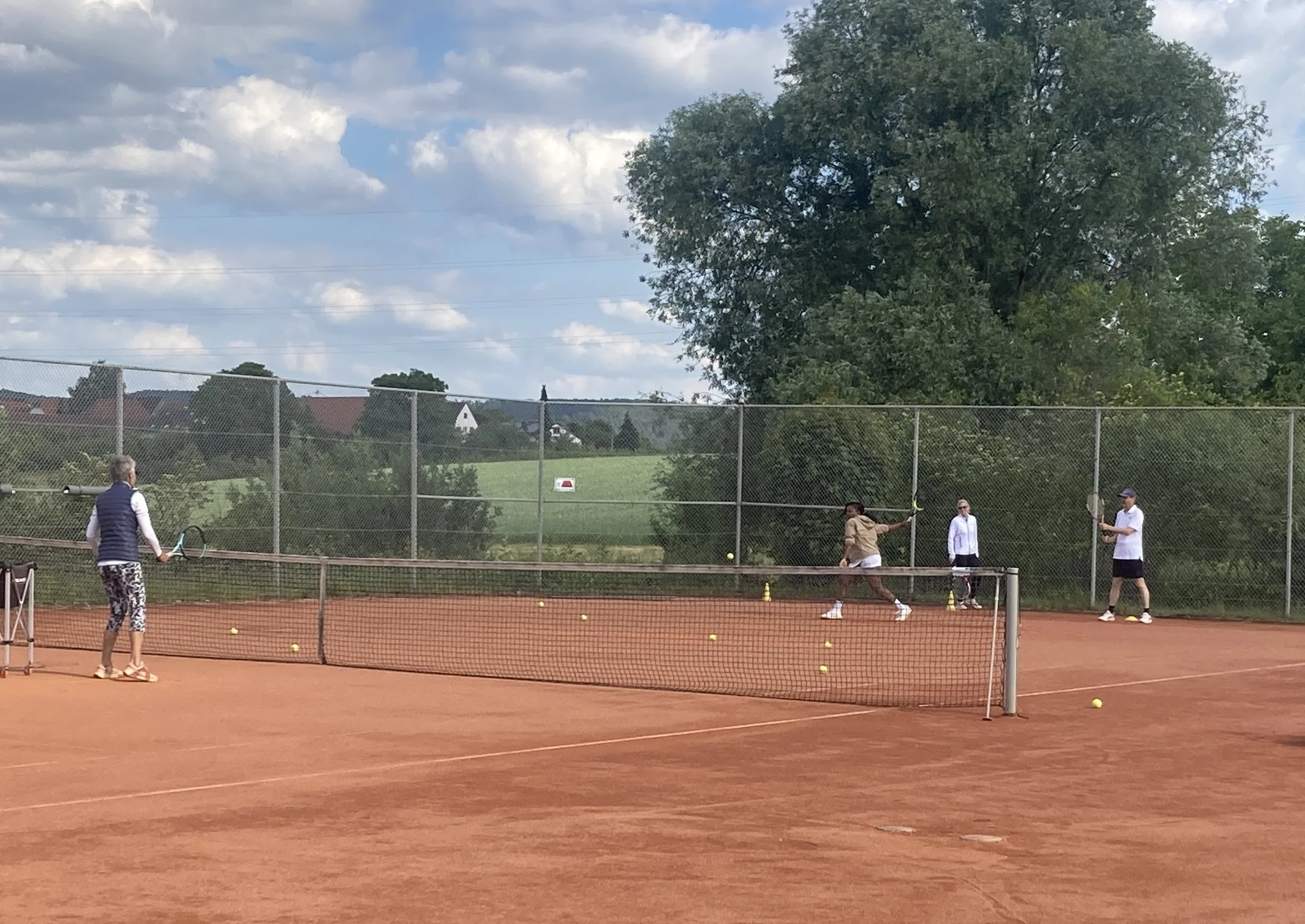 Vier Menschen spielen Tennis auf einem roten Tennisplatz im Freien, umgeben von Bäumen und einer grünen Landschaft, bei bewölktem Himmel.