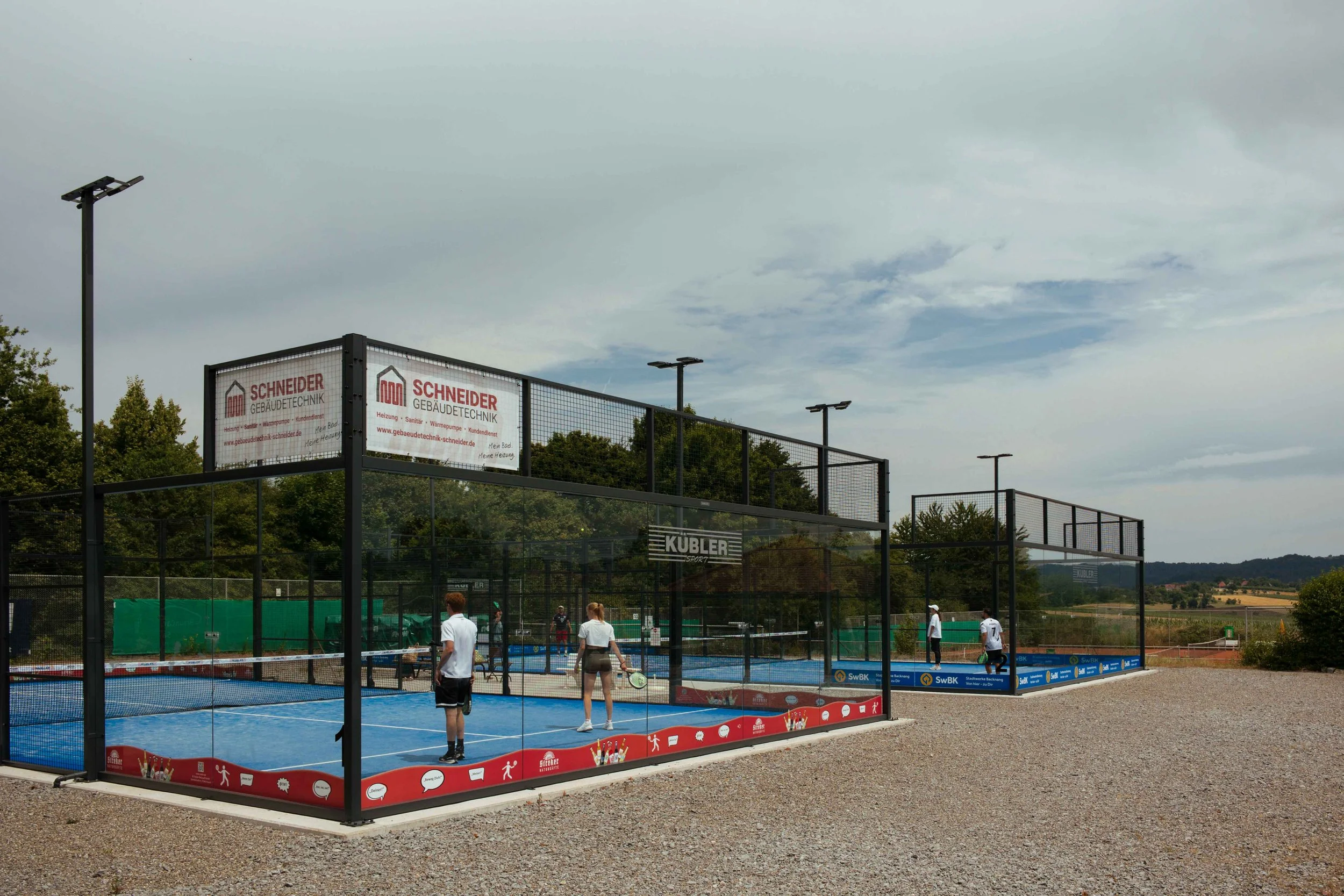 Zwei Padeltennisplätze im Freien, eingezäunt, mit Spielern, die auf dem Platz spielen, unter einem bewölkten Himmel, Landschaft im Hintergrund.